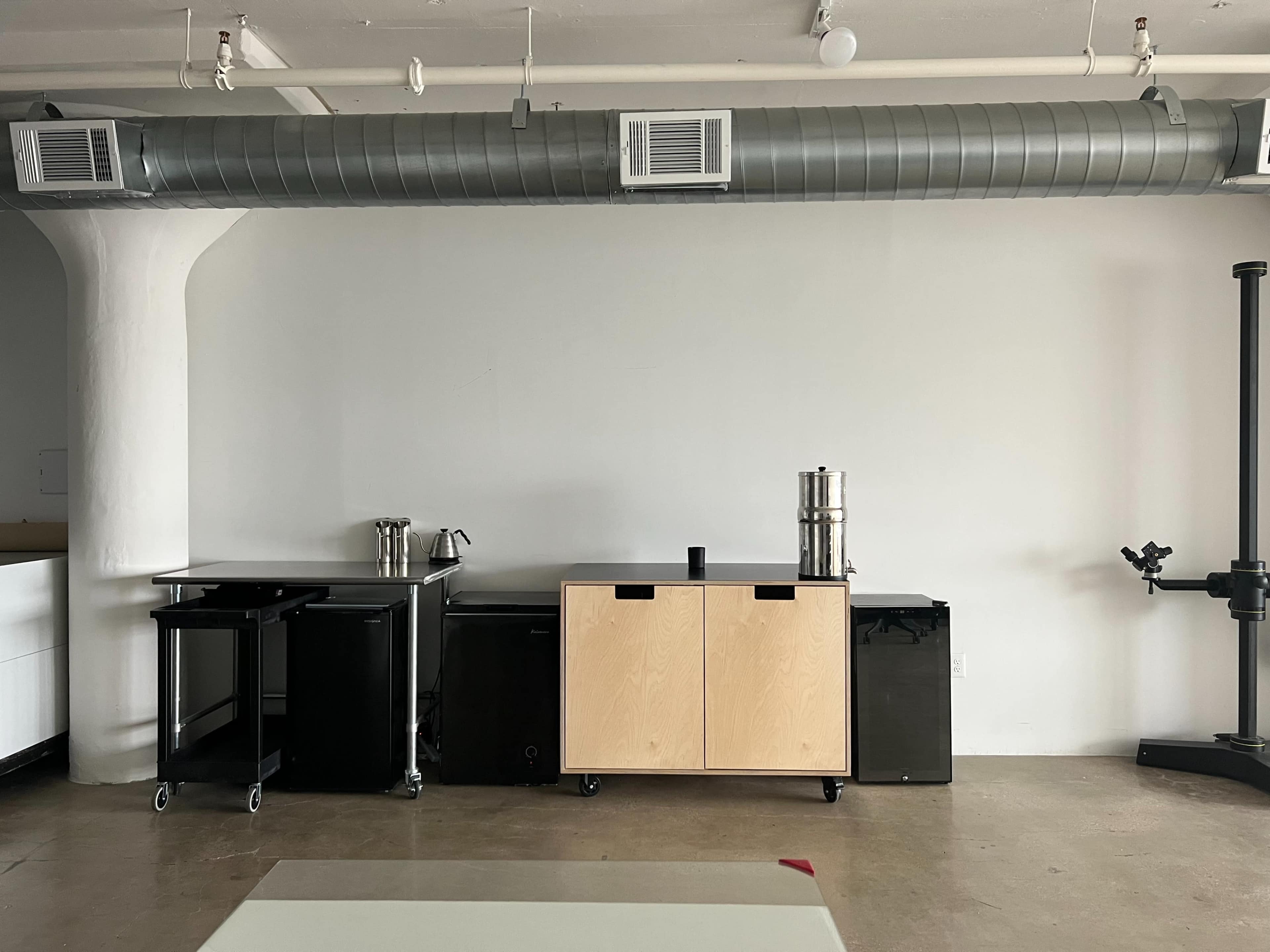 A minimalistic workspace with kitchen carts, a coffee urn, and a tall black cylindrical object against a plain white wall.