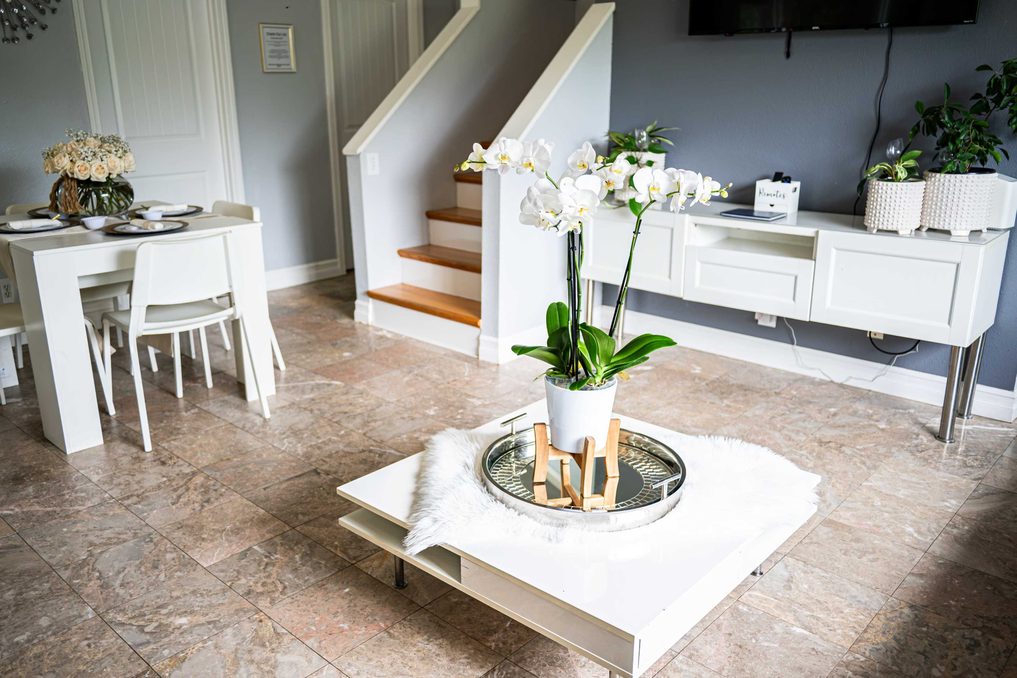 The image shows a modern living space featuring a coffee table with a white orchid centerpiece, a dining table with place settings, and a staircase in the background.