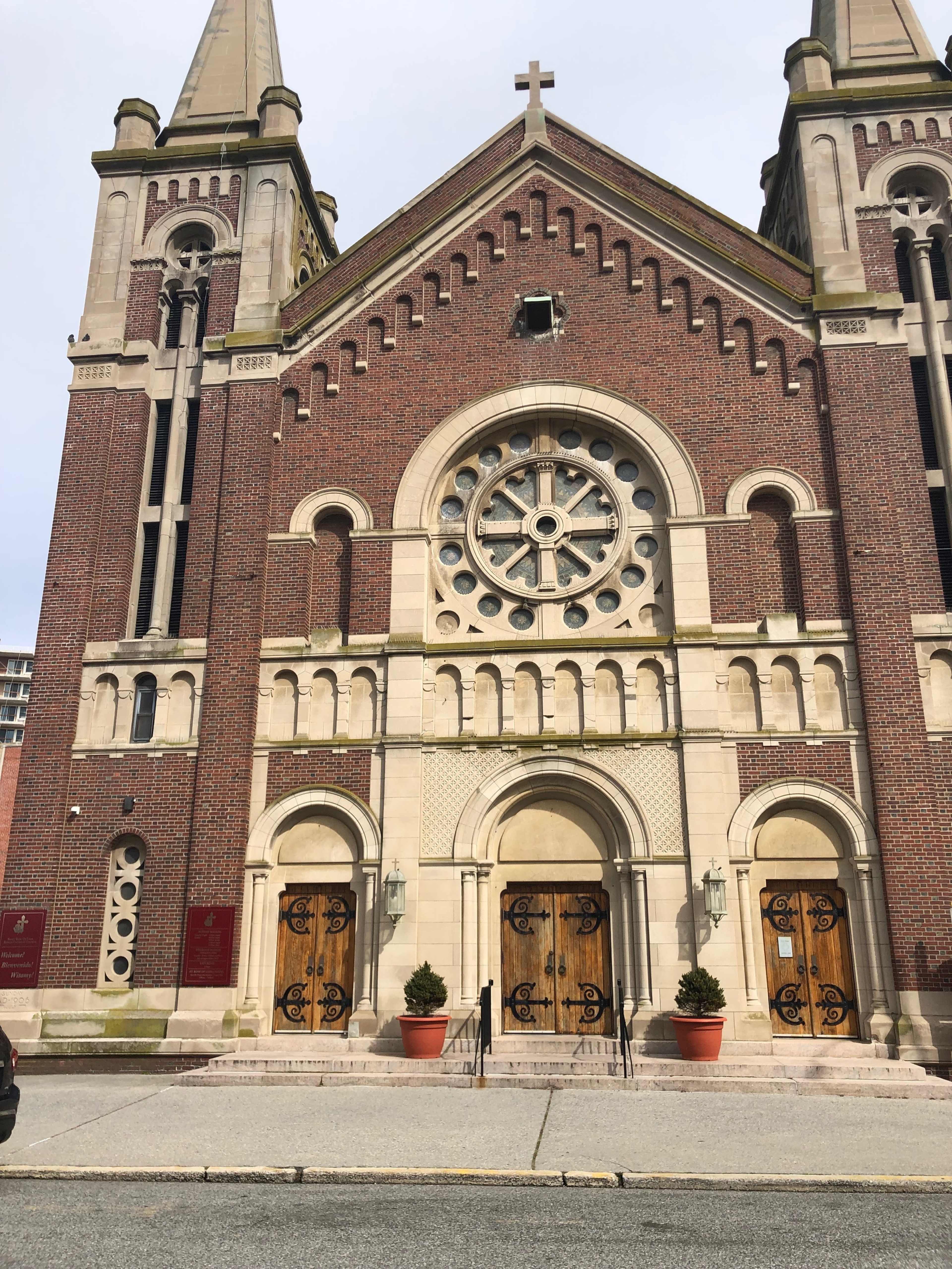 A brick church with two tall spires, a large rose window, and wooden double doors at the entrance.