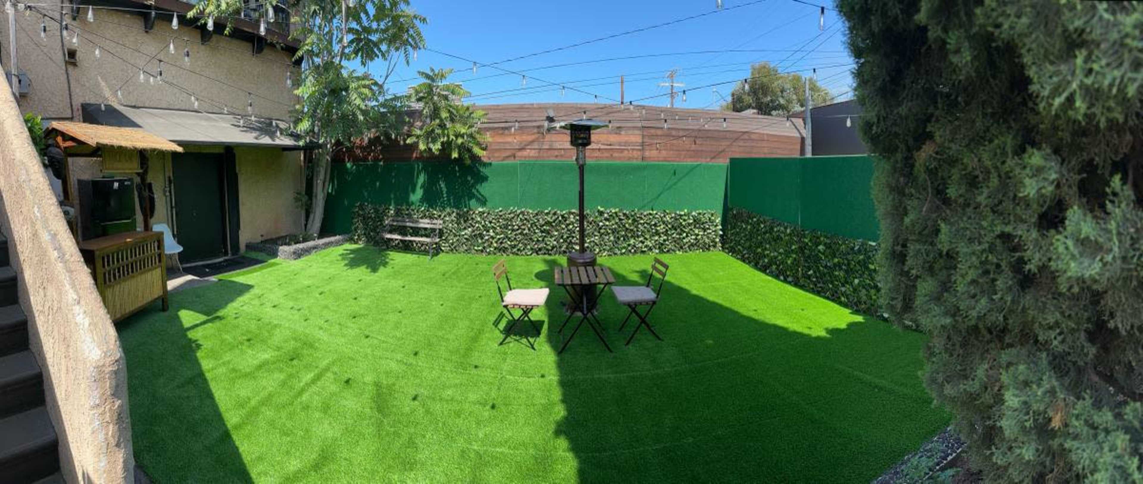 A panoramic view of a green artificial grass yard featuring a table with four chairs, surrounded by hedges and a tall heater.