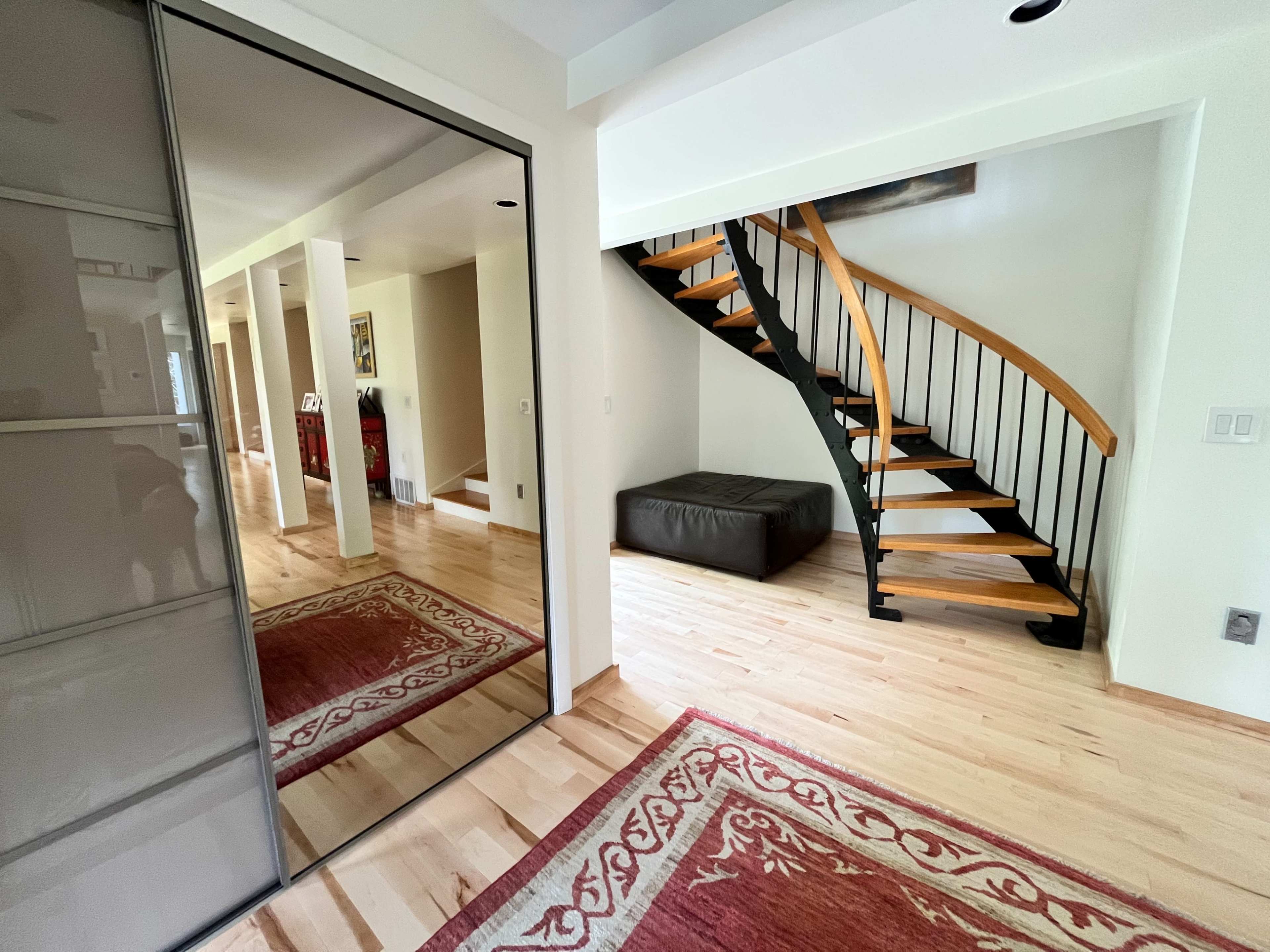 A hallway with a mirrored wardrobe, a wooden spiral staircase, and a leather ottoman positioned near a decorative rug.