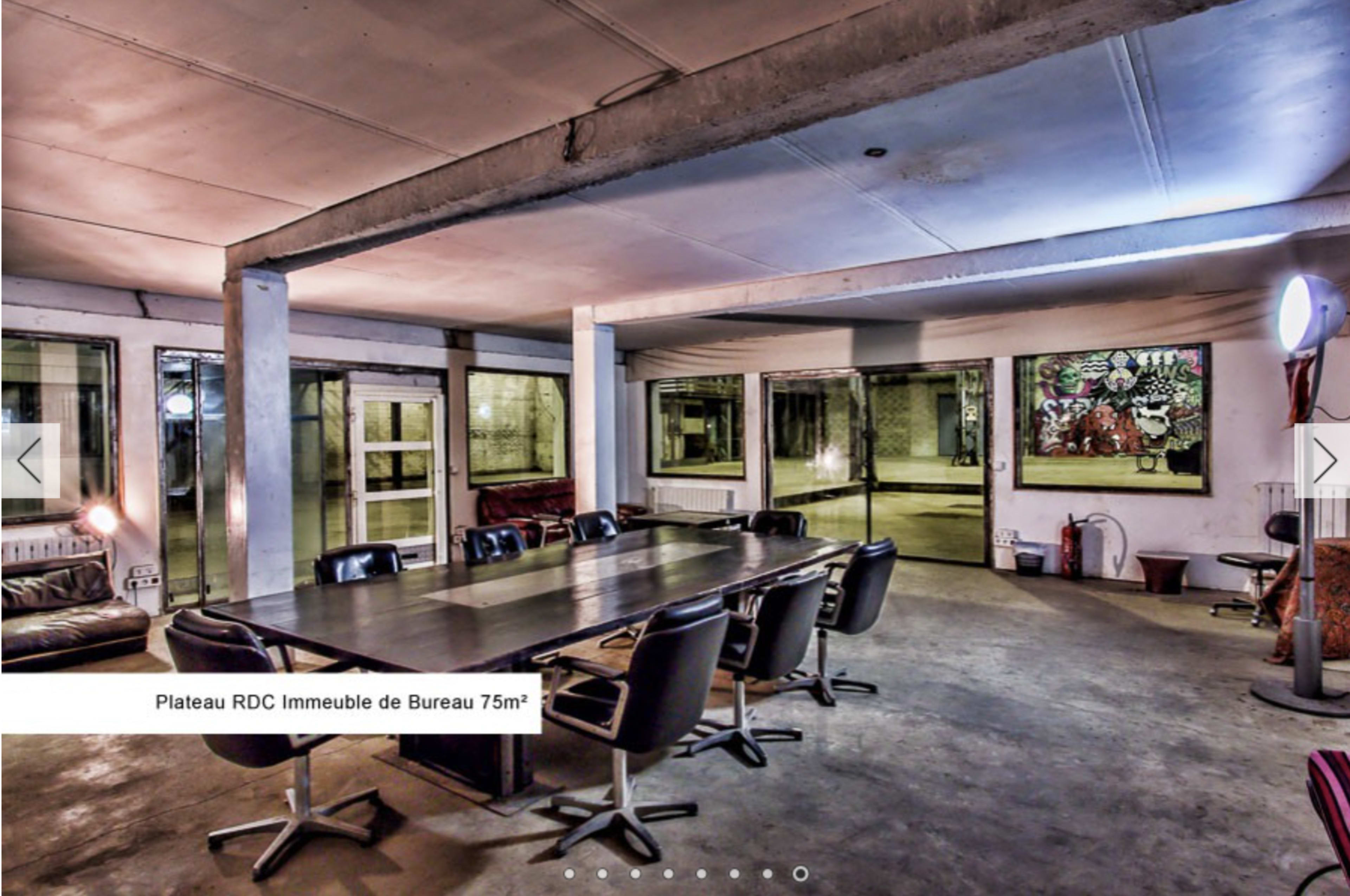 A modern office space with a large rectangular table surrounded by black chairs, concrete walls, and large windows.