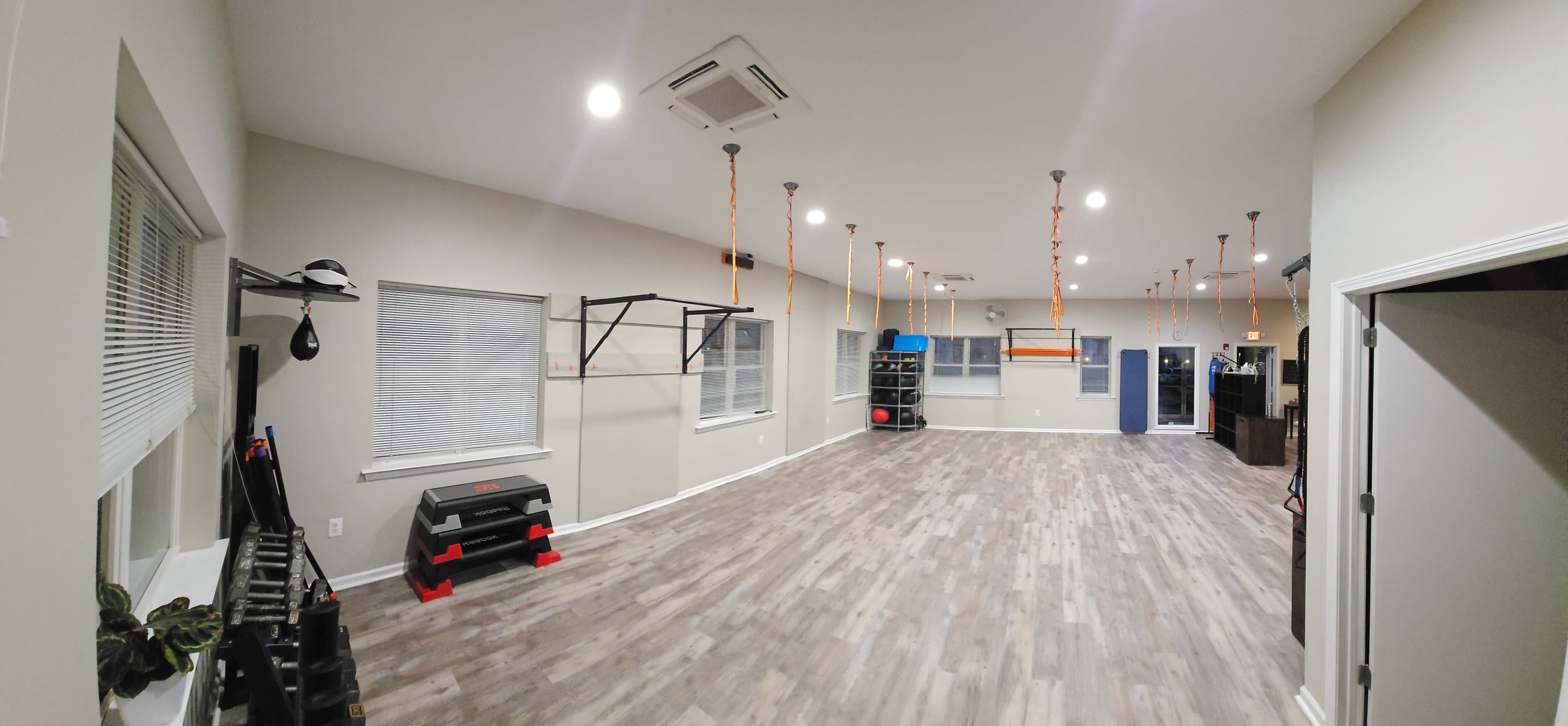 A spacious, well-lit gym with wooden flooring, various exercise equipment along the walls, and suspended ropes hanging from the ceiling.