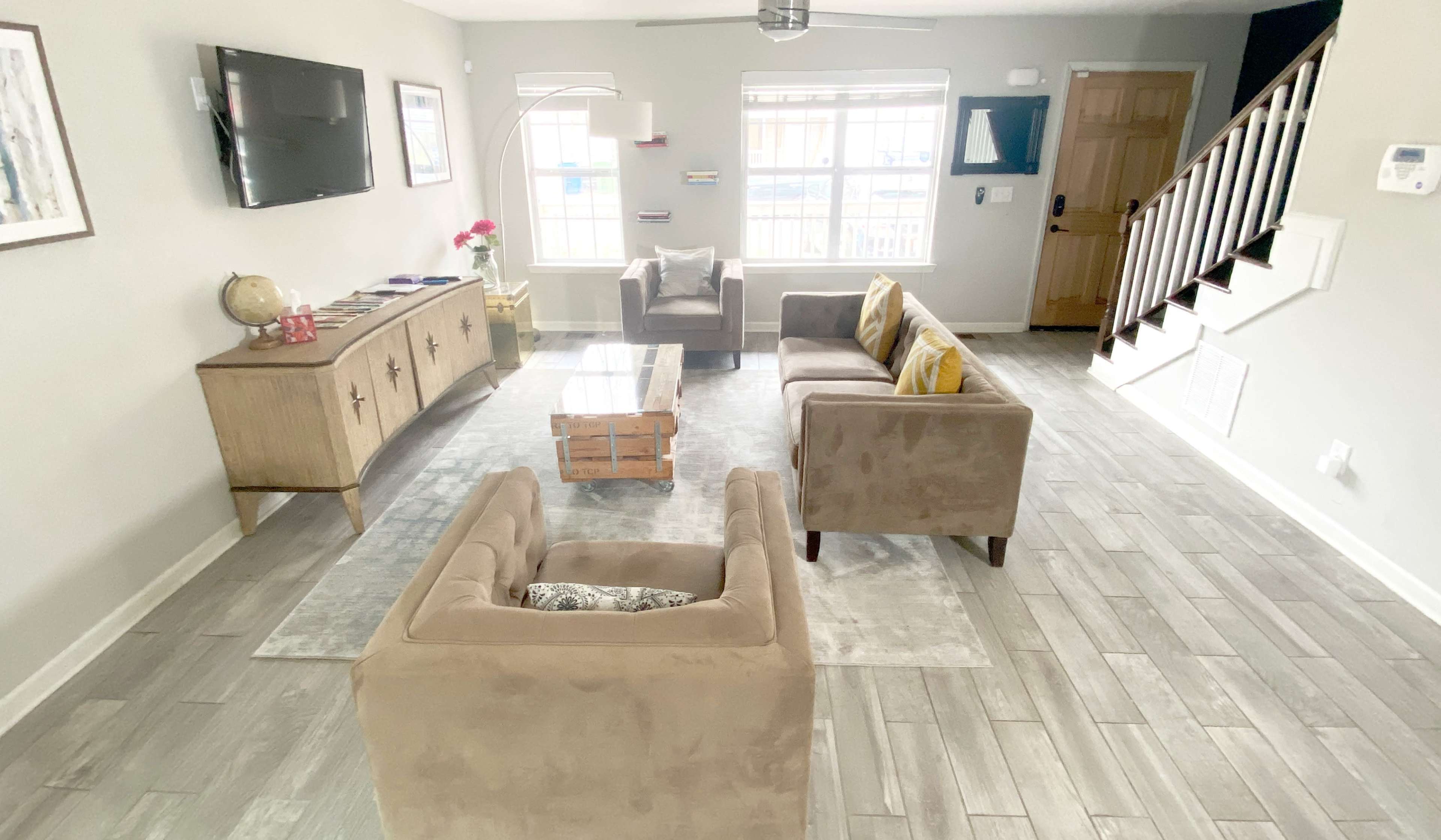 The image shows a living room with two brown chairs, a coffee table, a beige sofa, a television mounted on the wall, and a staircase in the background.