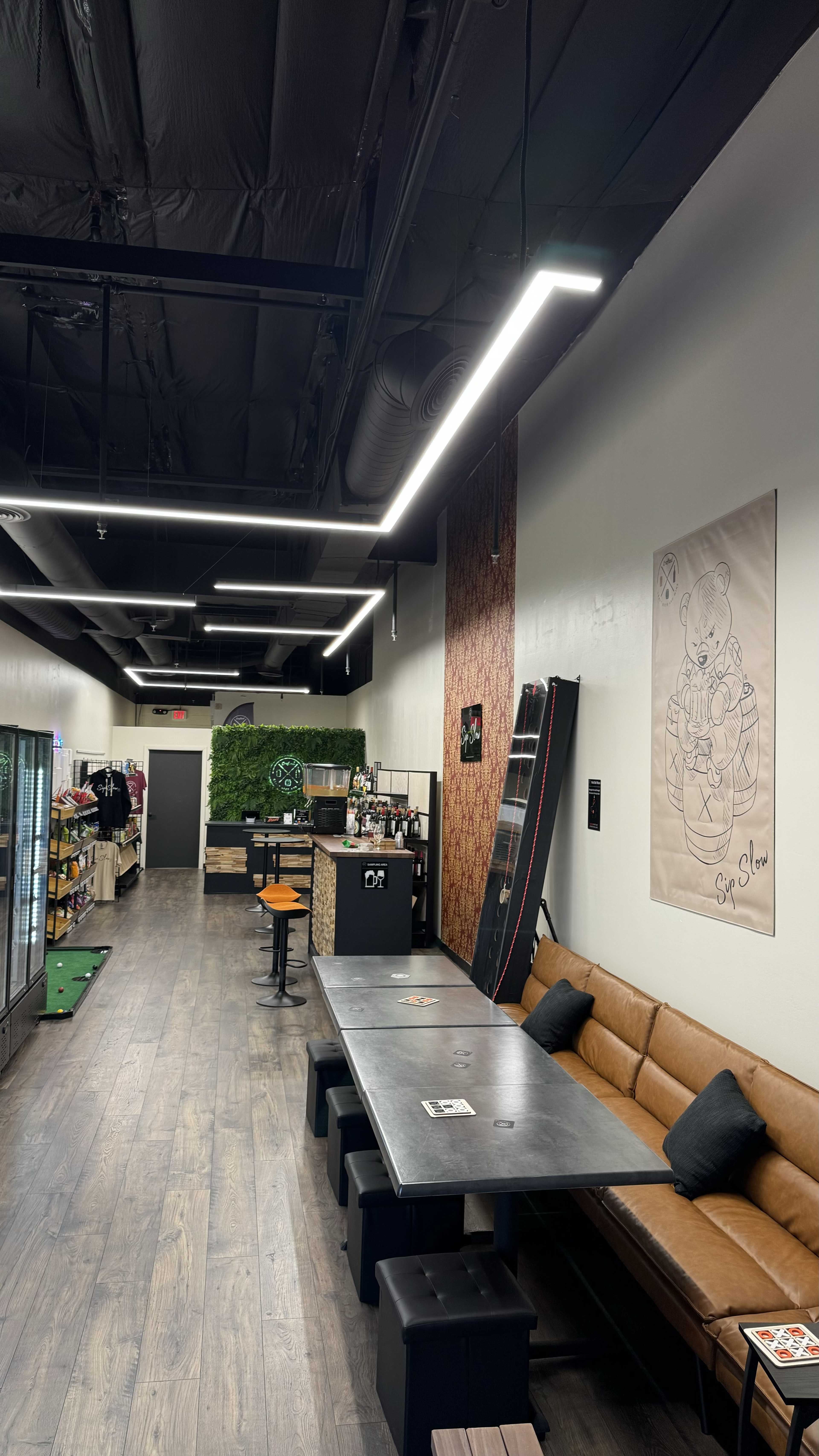 The image shows an interior of a modern café or lounge area with a long table, a couch, and a bar-like seating arrangement along one wall, alongside a shelf displaying drinks.