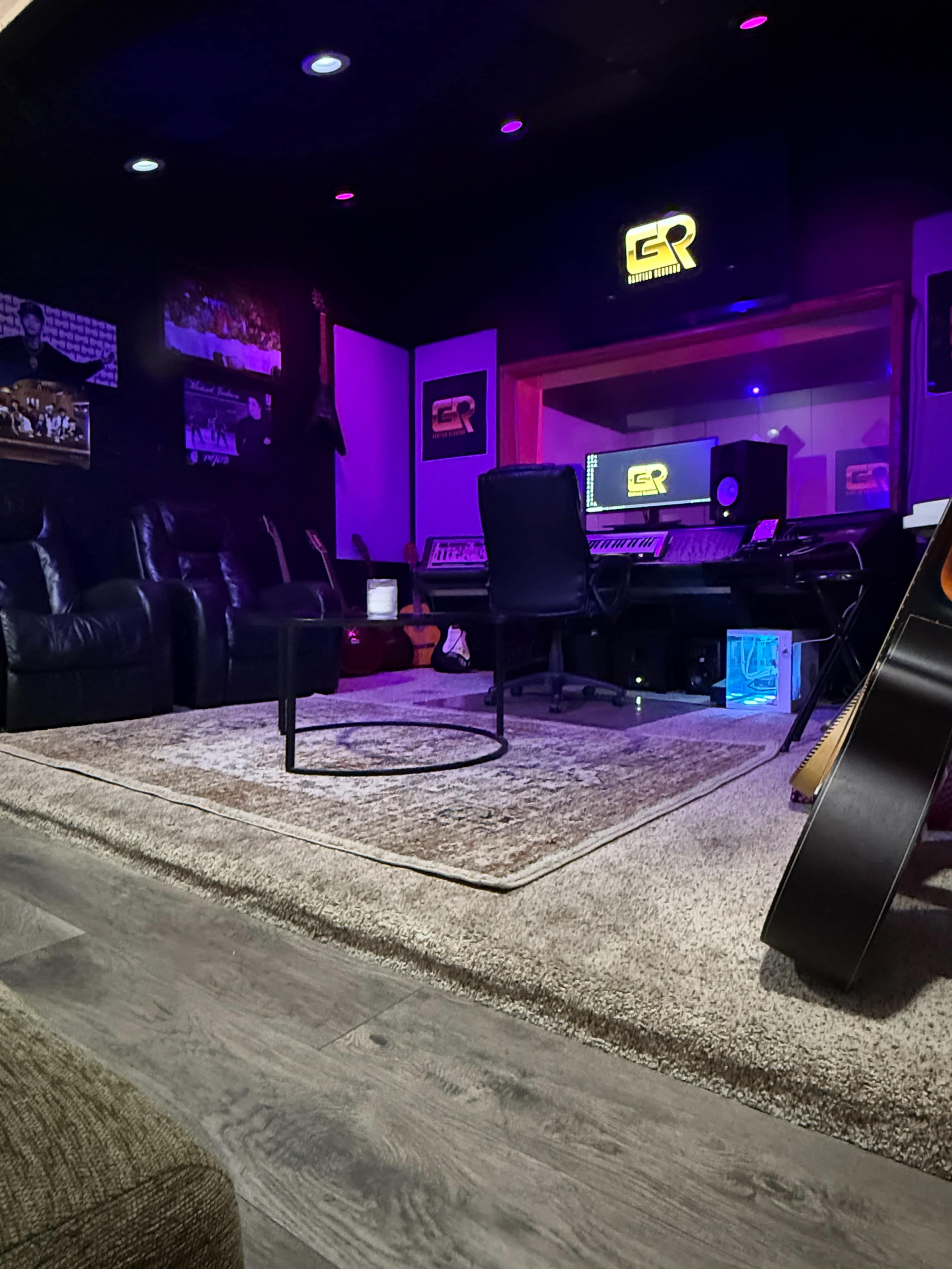 The image shows a modern music studio featuring a mixing console, leather seating, and guitars, illuminated by purple and blue lights.