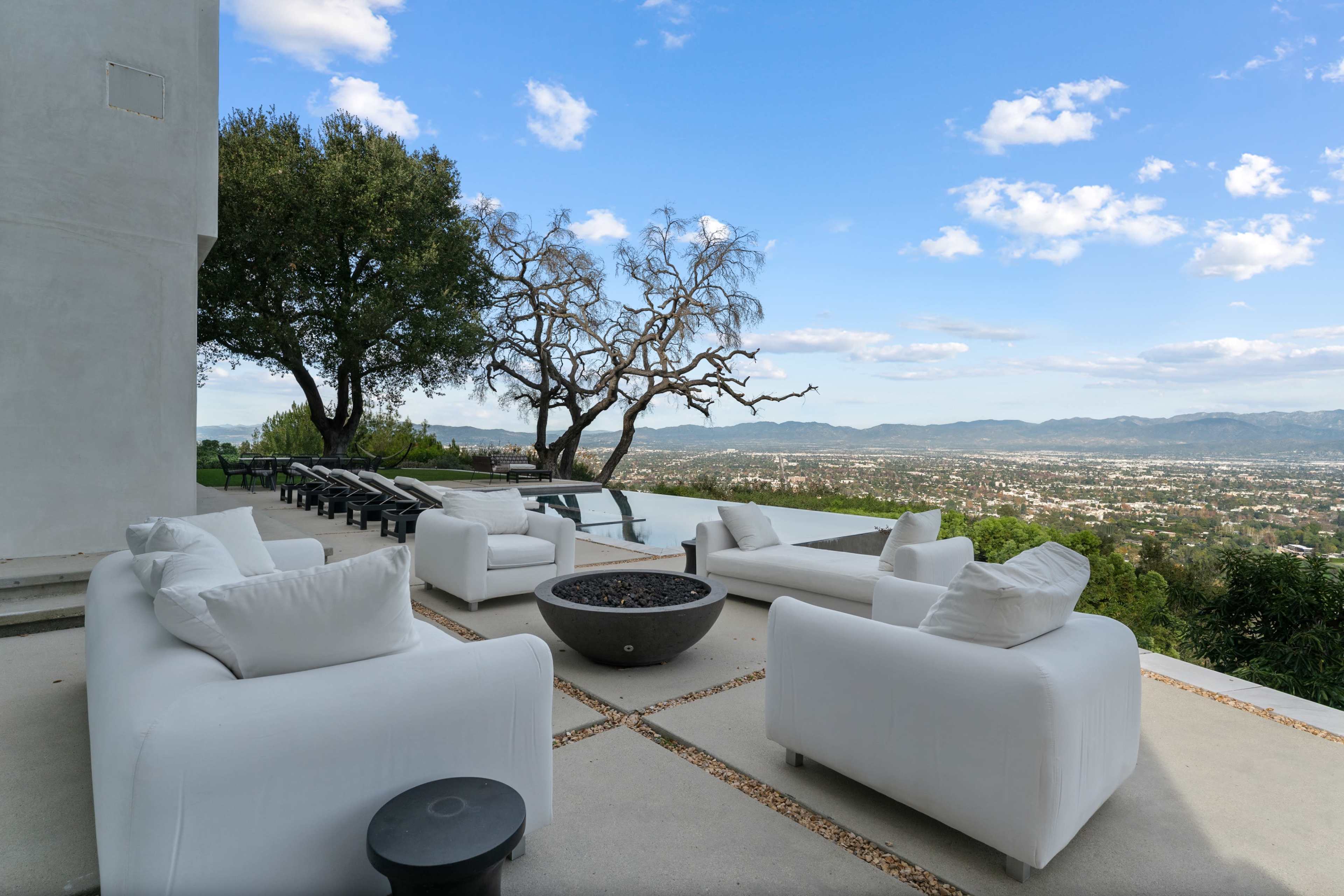 A modern outdoor patio features white furniture and a fire pit, overlooking a valley with mountains in the background under a partly cloudy sky.