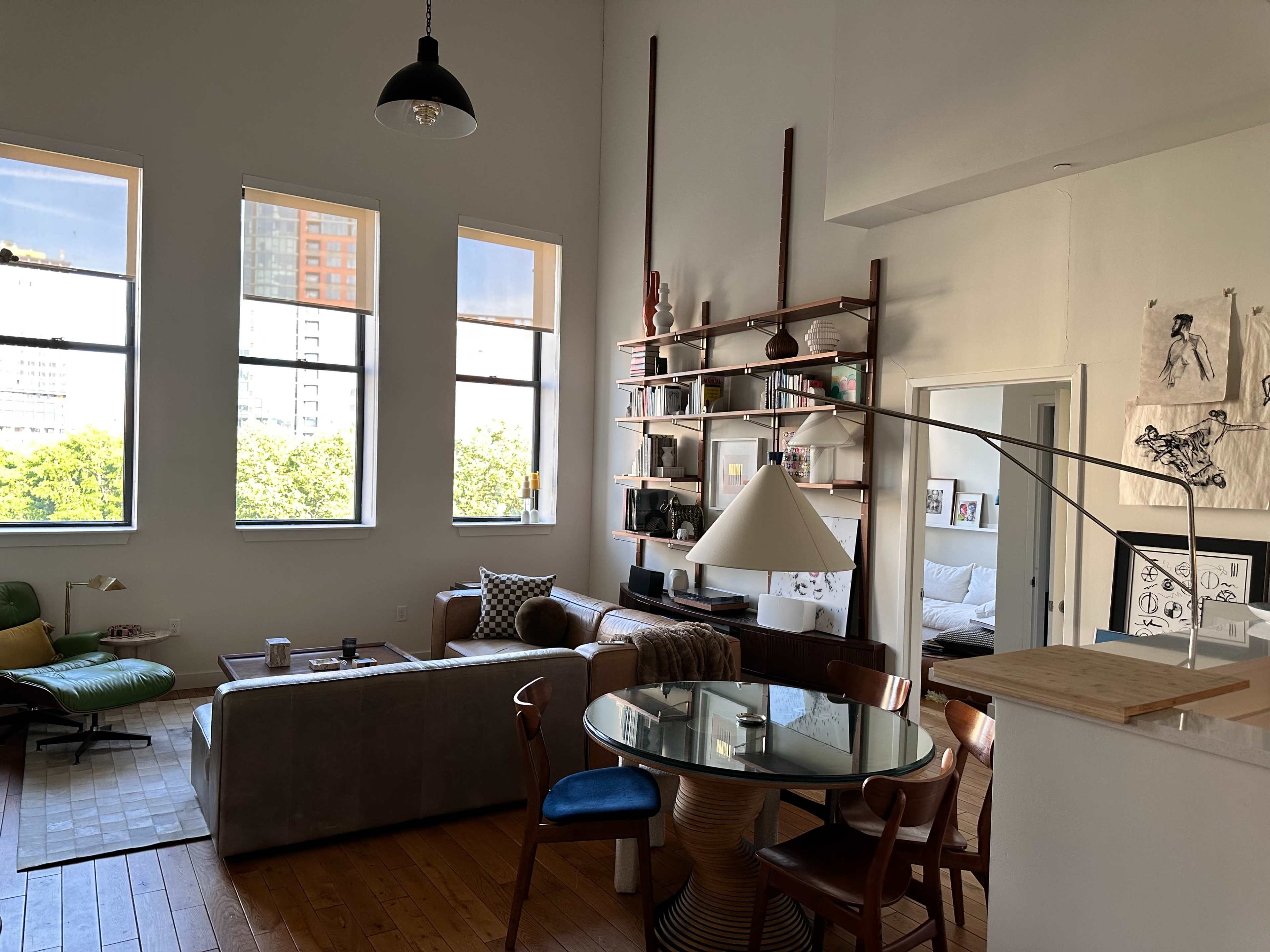 The image shows a modern apartment interior with a living area featuring large windows, a sectional sofa, a dining table, and a bookshelf.