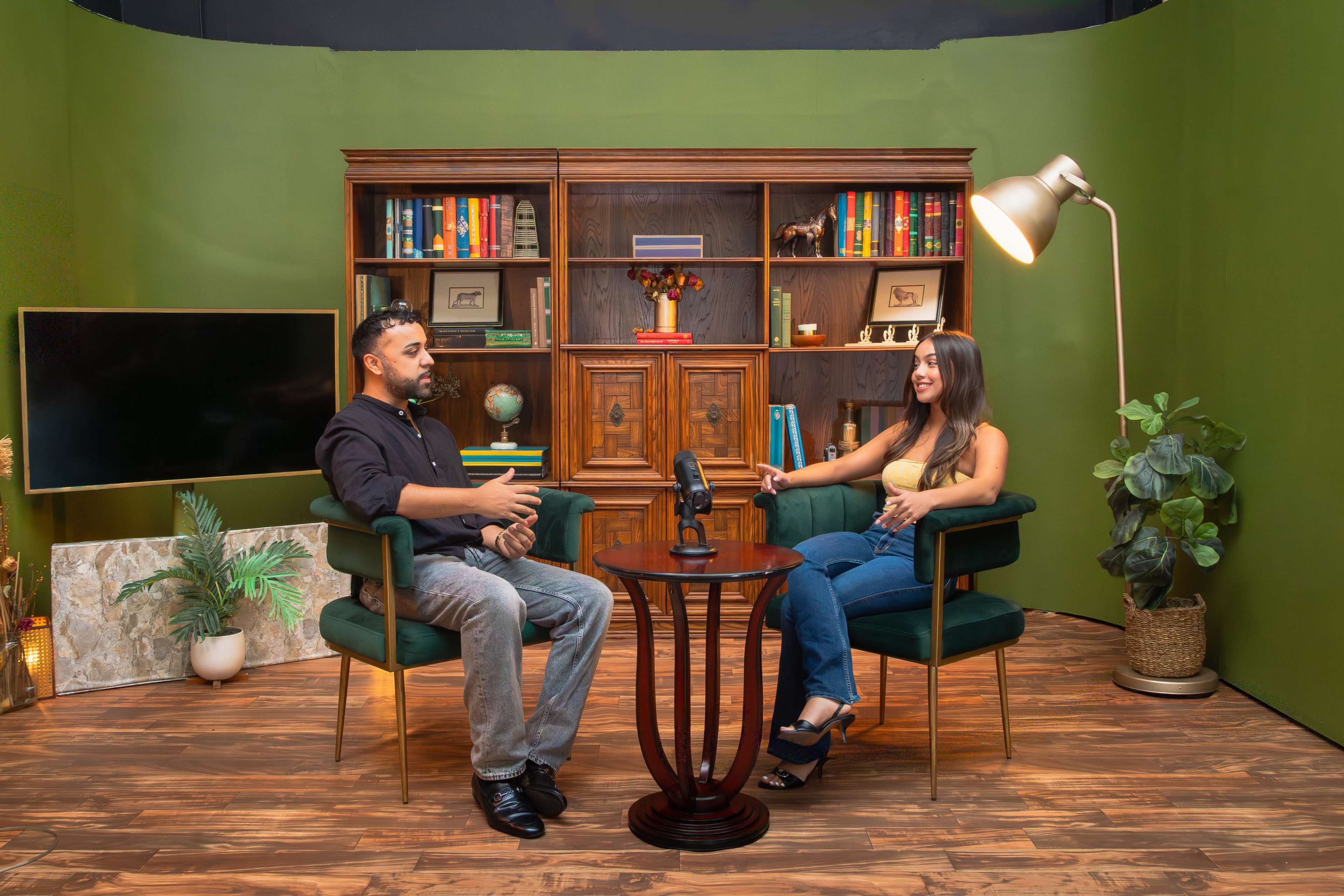 A man and a woman are seated in a well-decorated studio with green walls, engaged in conversation near a microphone on a small table.
