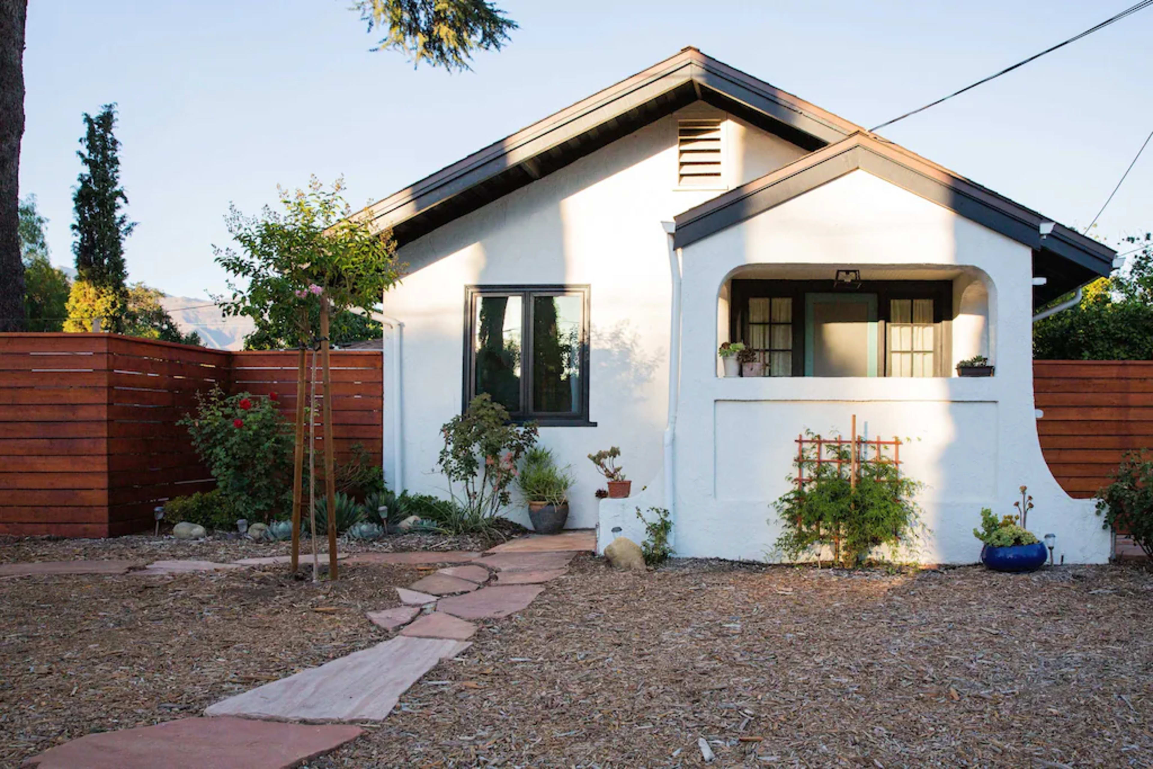 A white cottage-style house with a paved pathway leads to a front porch, surrounded by a garden and wooden fence.