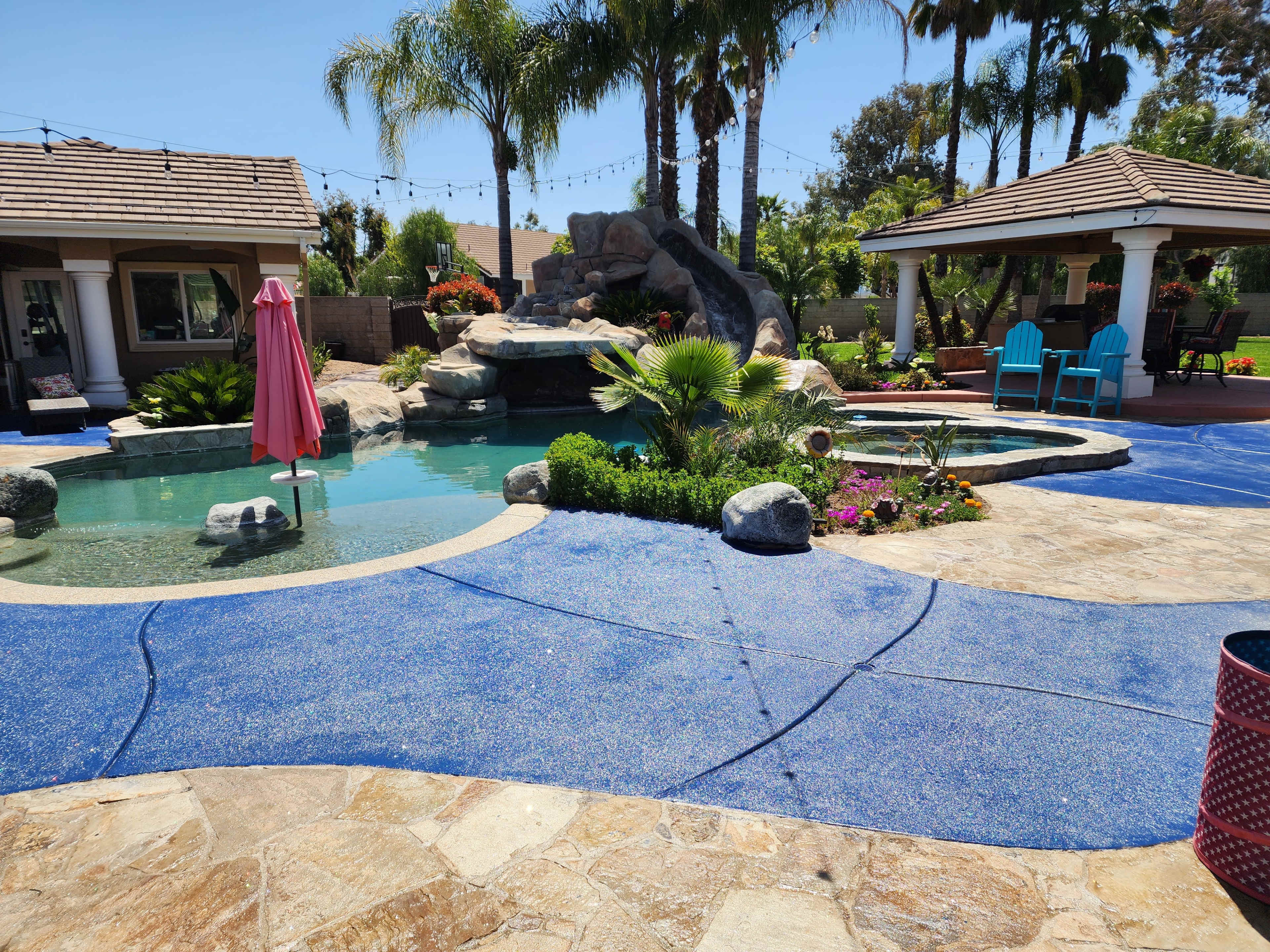 A backyard pool area featuring a rock waterfall, tropical landscaping, and a cabana with lounge chairs.
