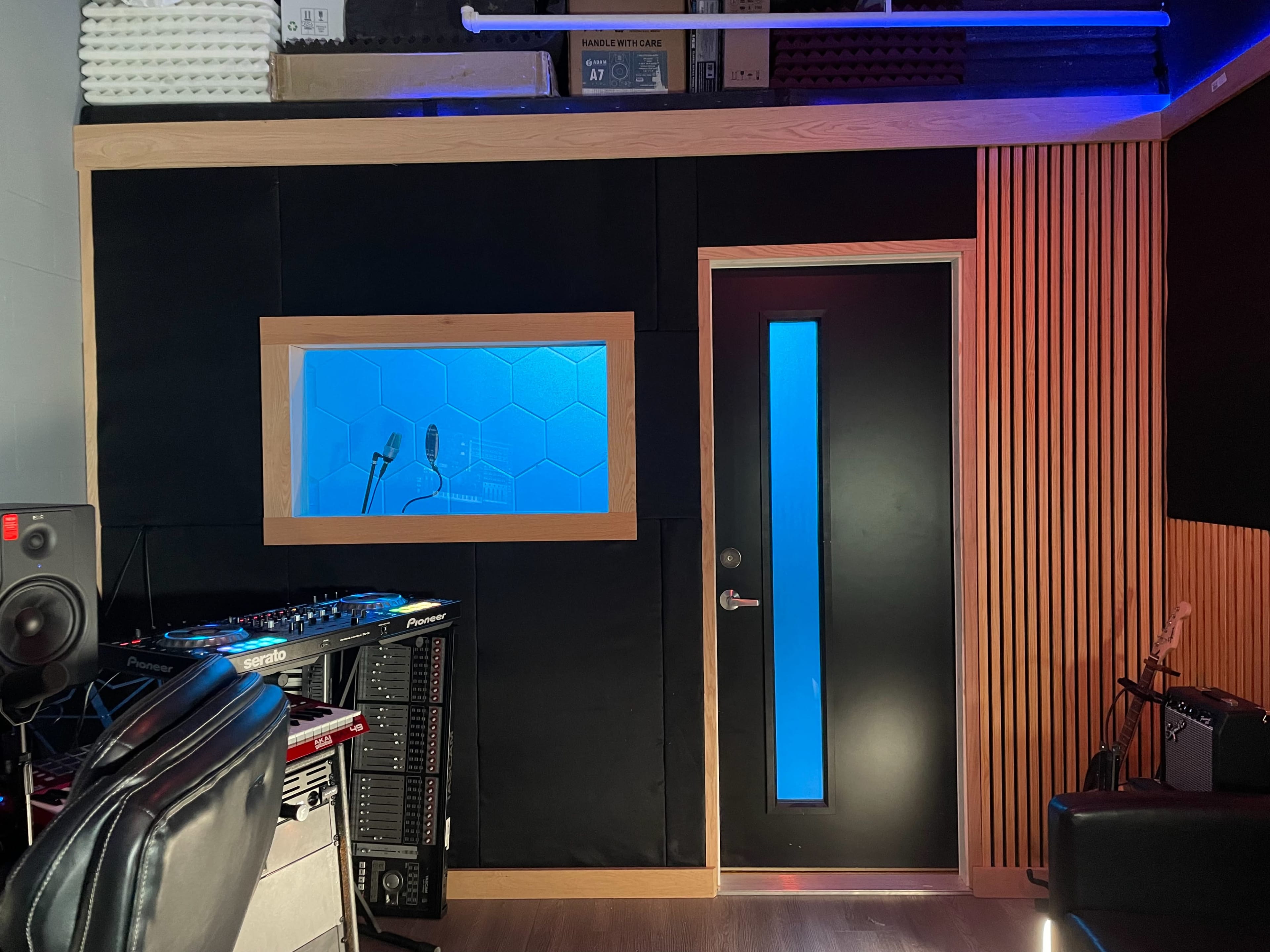 The image shows a sound studio featuring a control area with a door and a window that displays a blue backlight, along with soundproof walls and recording equipment.