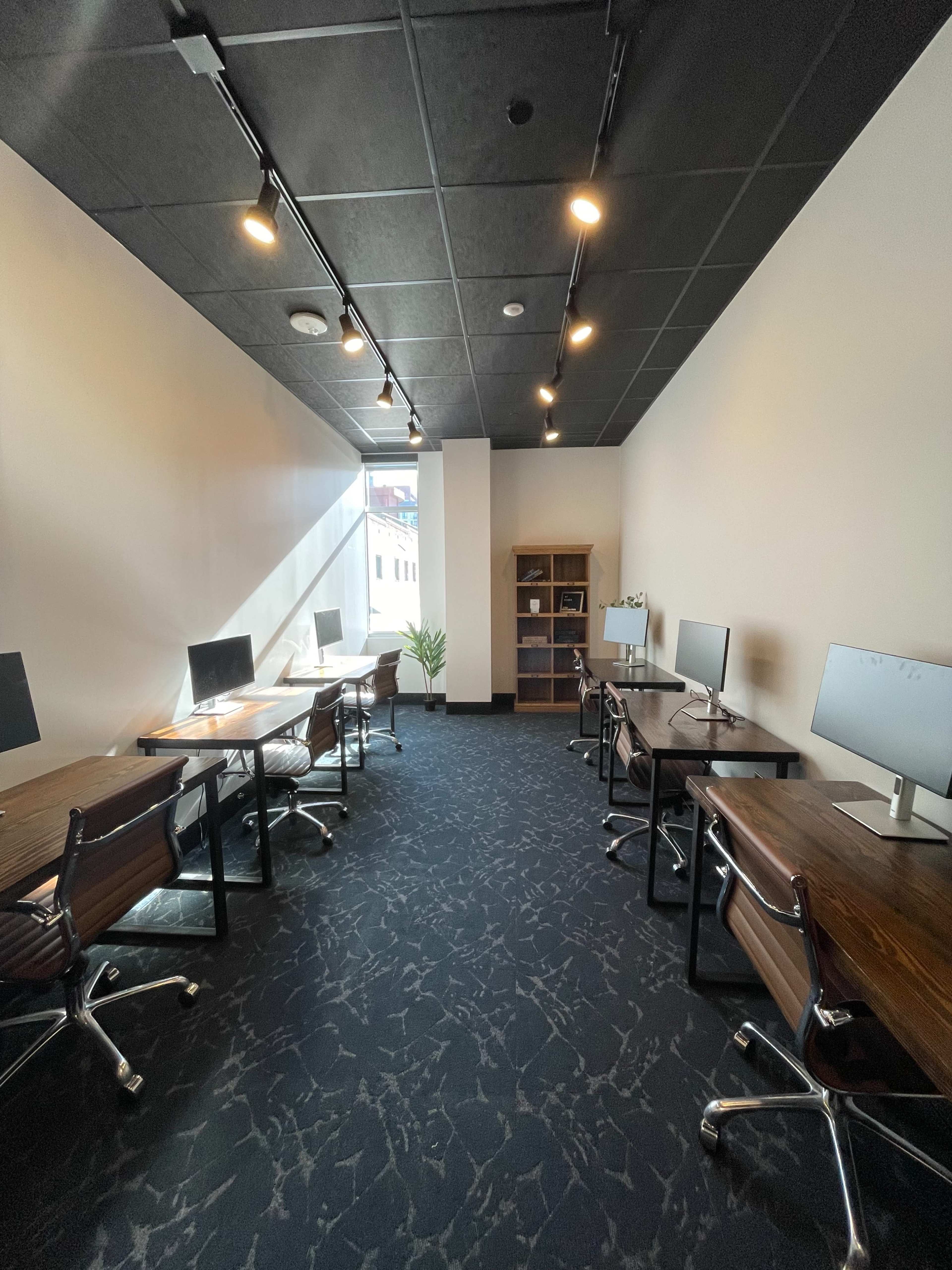 The image shows a small office space with several desks and computers arranged along the walls, featuring a bookshelf and a window providing natural light.