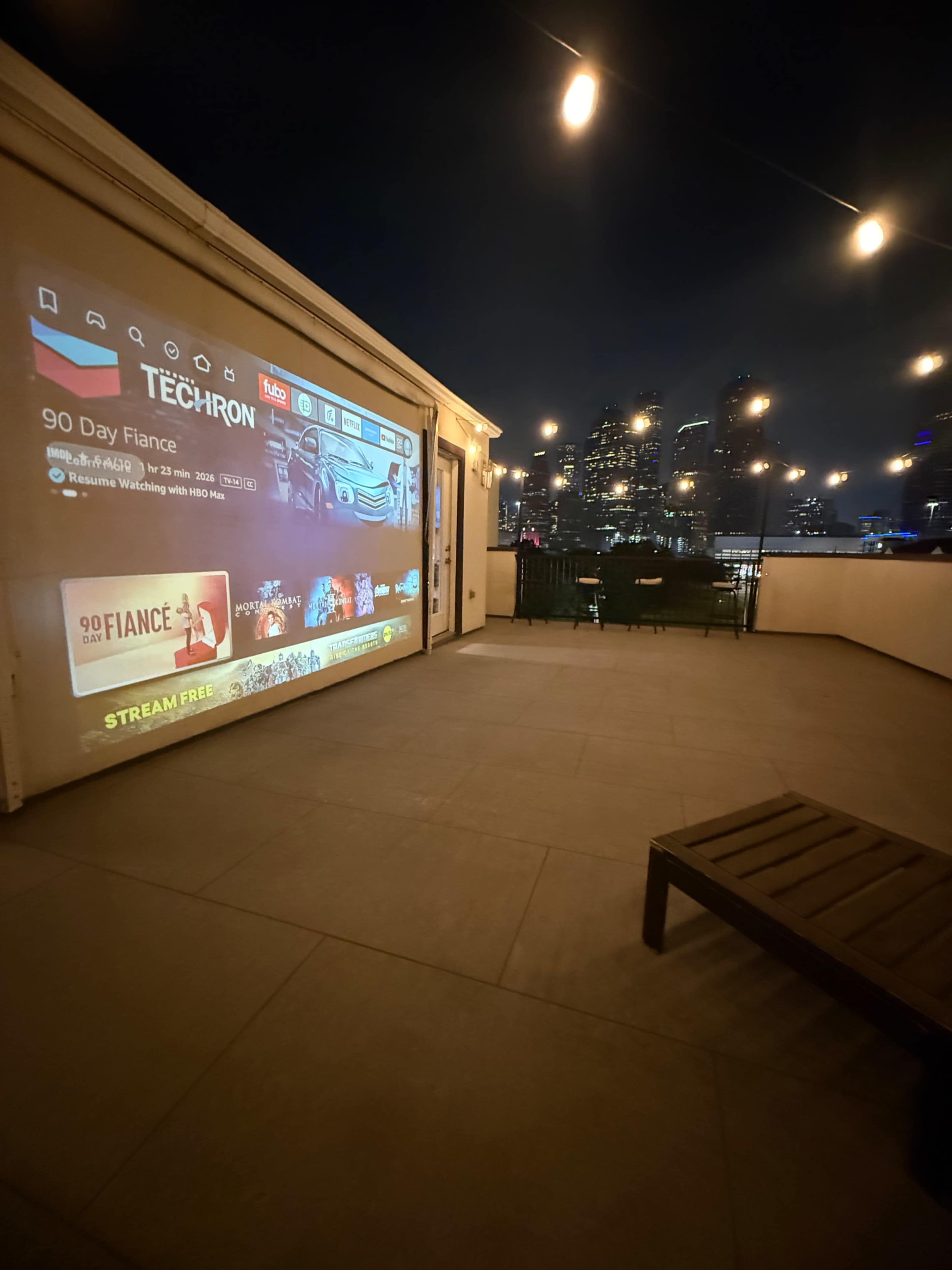A large outdoor projection screen displays a streaming service interface on a modern terrace at night, overlooking a city skyline.