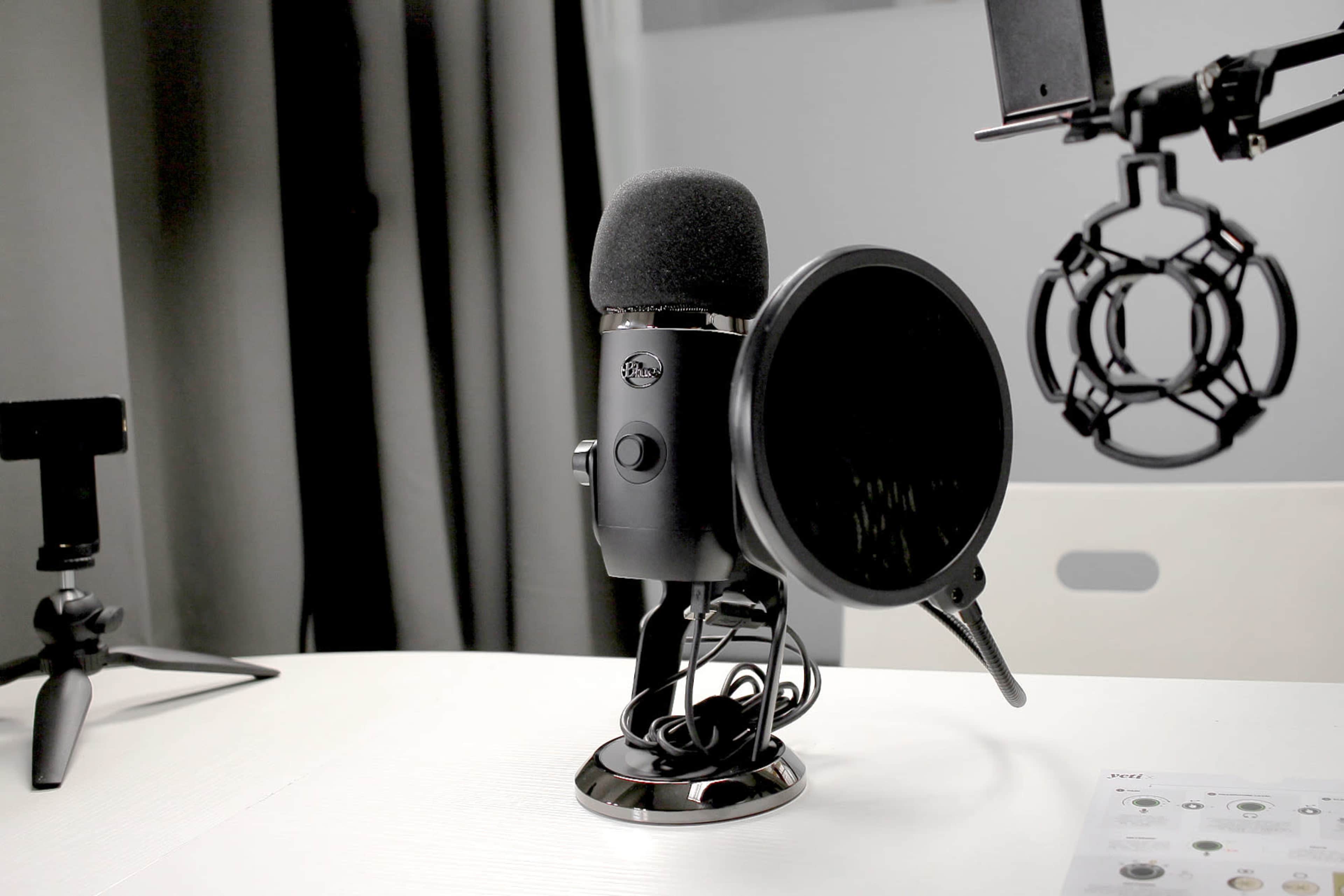 A black microphone with a pop filter is positioned on a white desk, alongside a tripod and an audio interface.