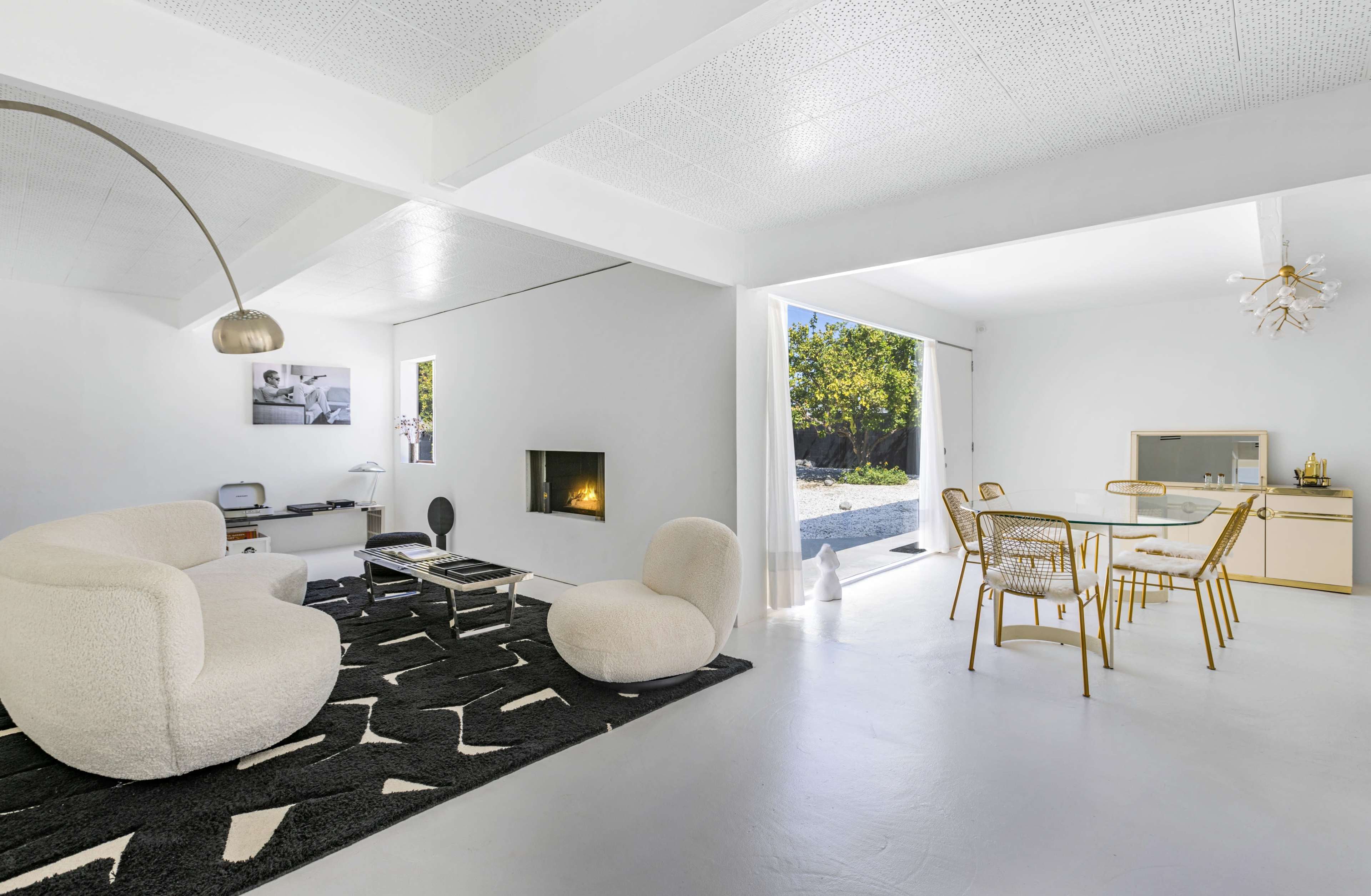 A modern living space features a white color scheme, a cozy seating area, a dining table with gold accents, and large windows opening to an outdoor garden.
