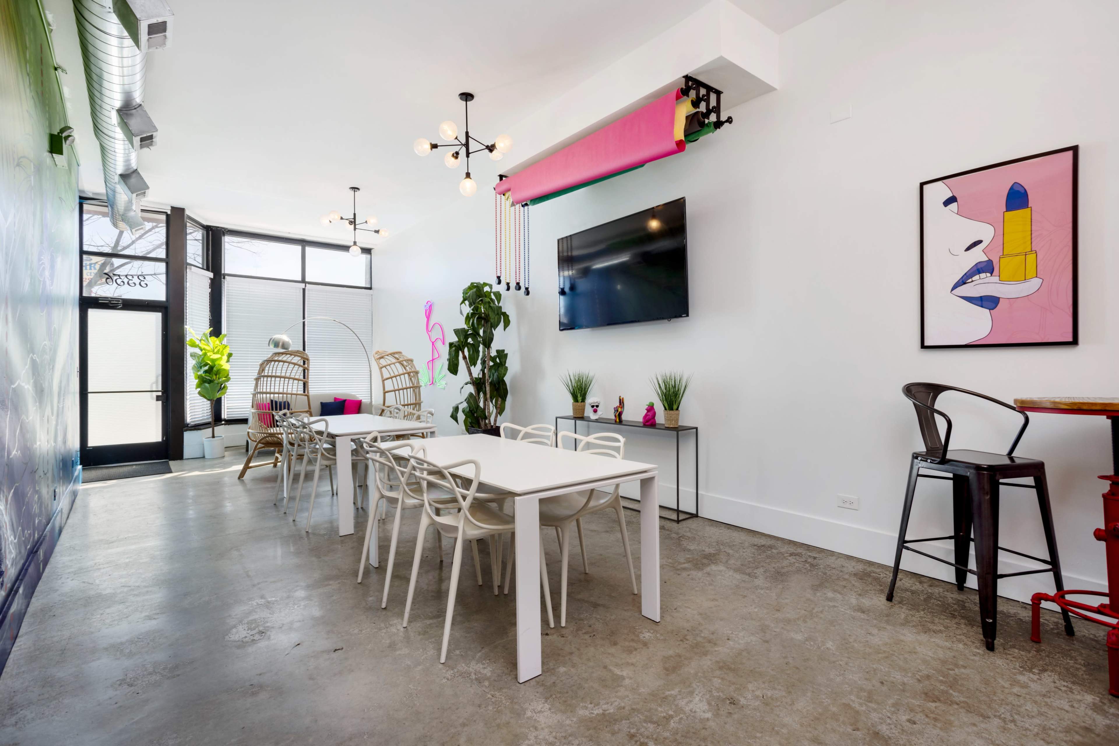 The image shows a modern interior space featuring a large table with white chairs, a wall-mounted television, and decorative plants, alongside colorful artwork and various lighting fixtures.