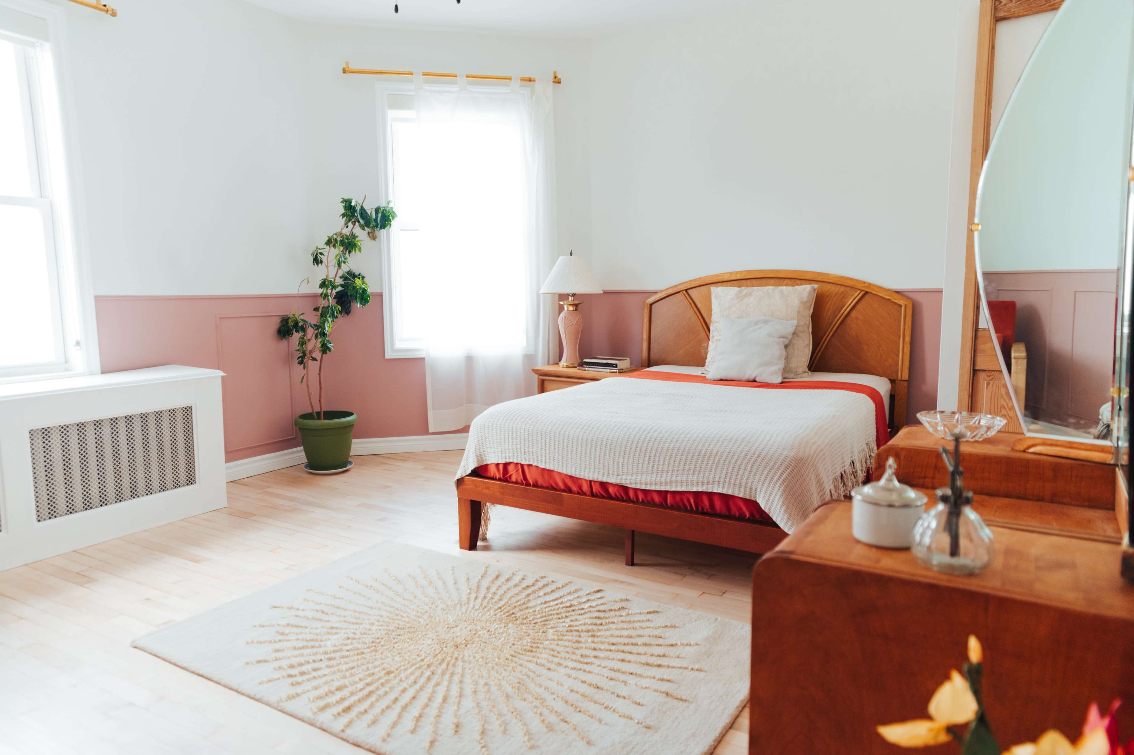 A bright bedroom features a bed with a white and red coverlet, a mirror, a potted plant, and a lamp beside the window.