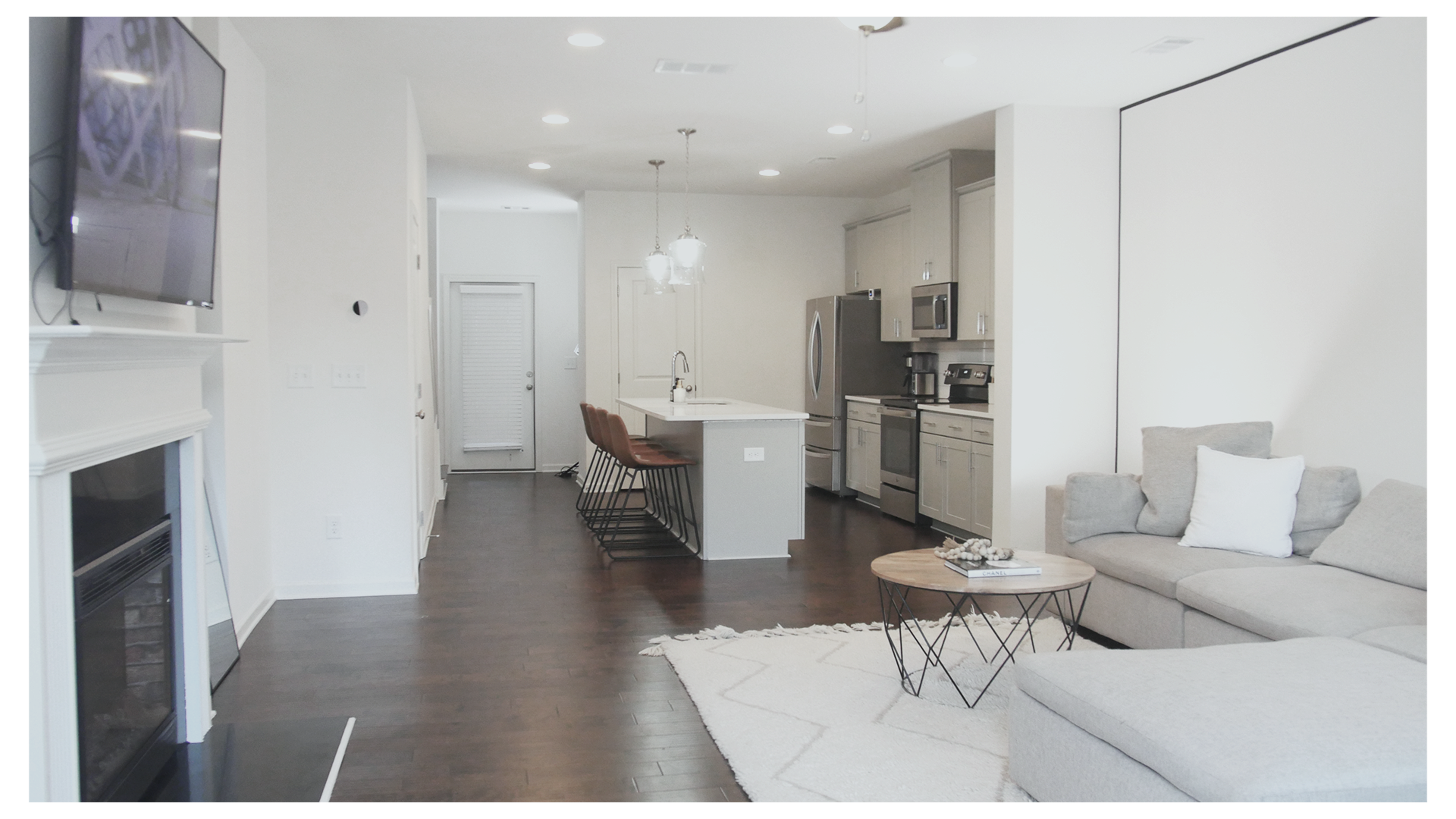 The image shows a modern open-plan living space featuring a kitchen with a large island, bar stools, a dining area, and a cozy seating area with a gray sectional sofa and a round coffee table.