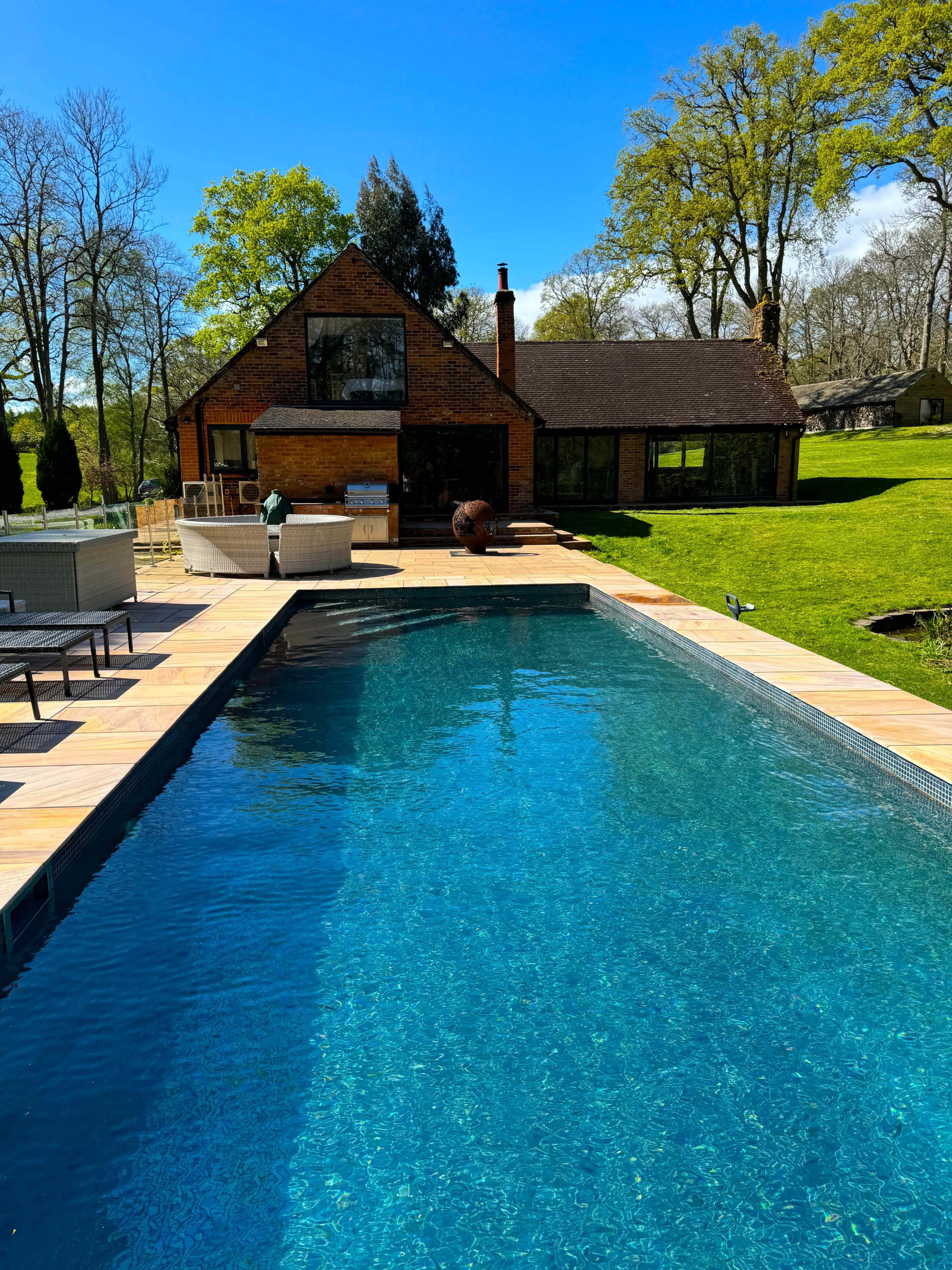 A clear blue swimming pool is situated in front of a modern brick house surrounded by lush greenery and trees under a bright blue sky.