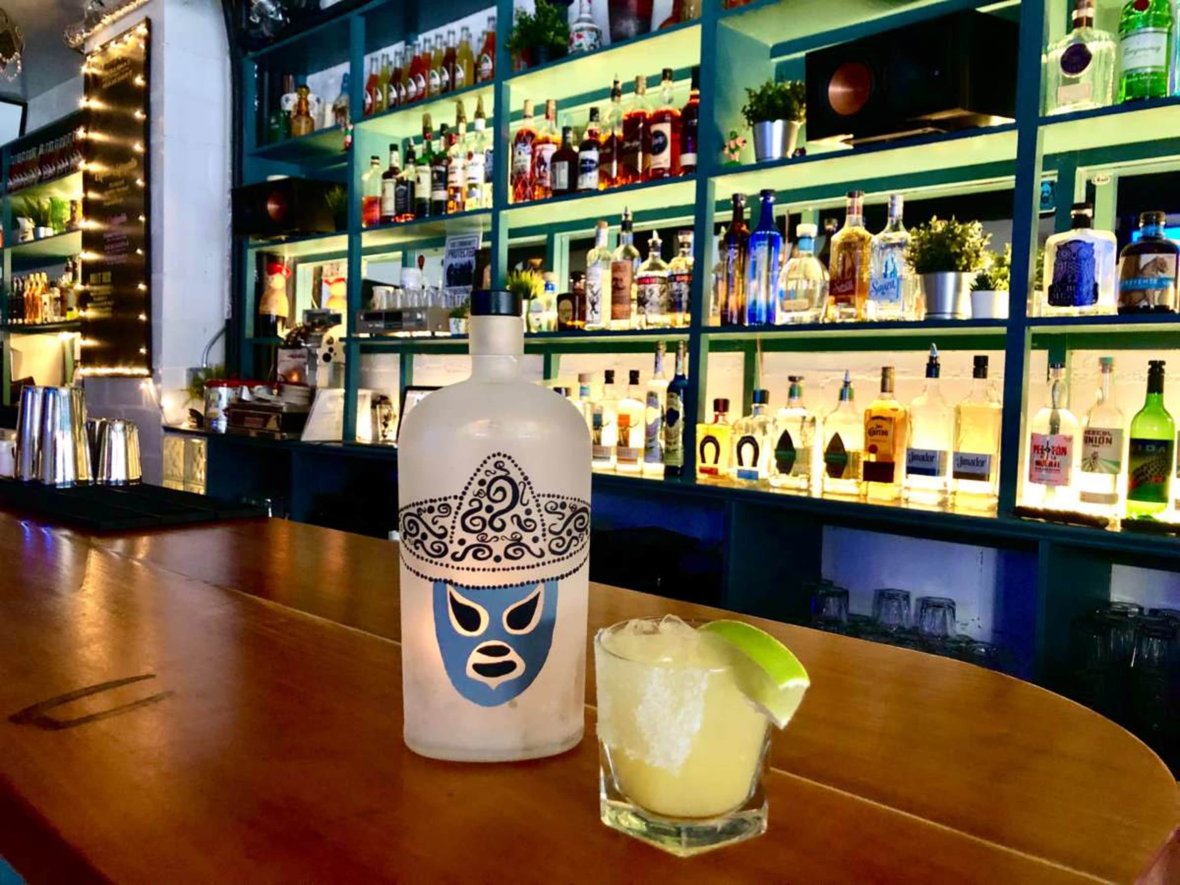 A bottle with a luchador mask design stands next to a glass of margarita on a wooden bar, with shelves of colorful liquor bottles in the background.