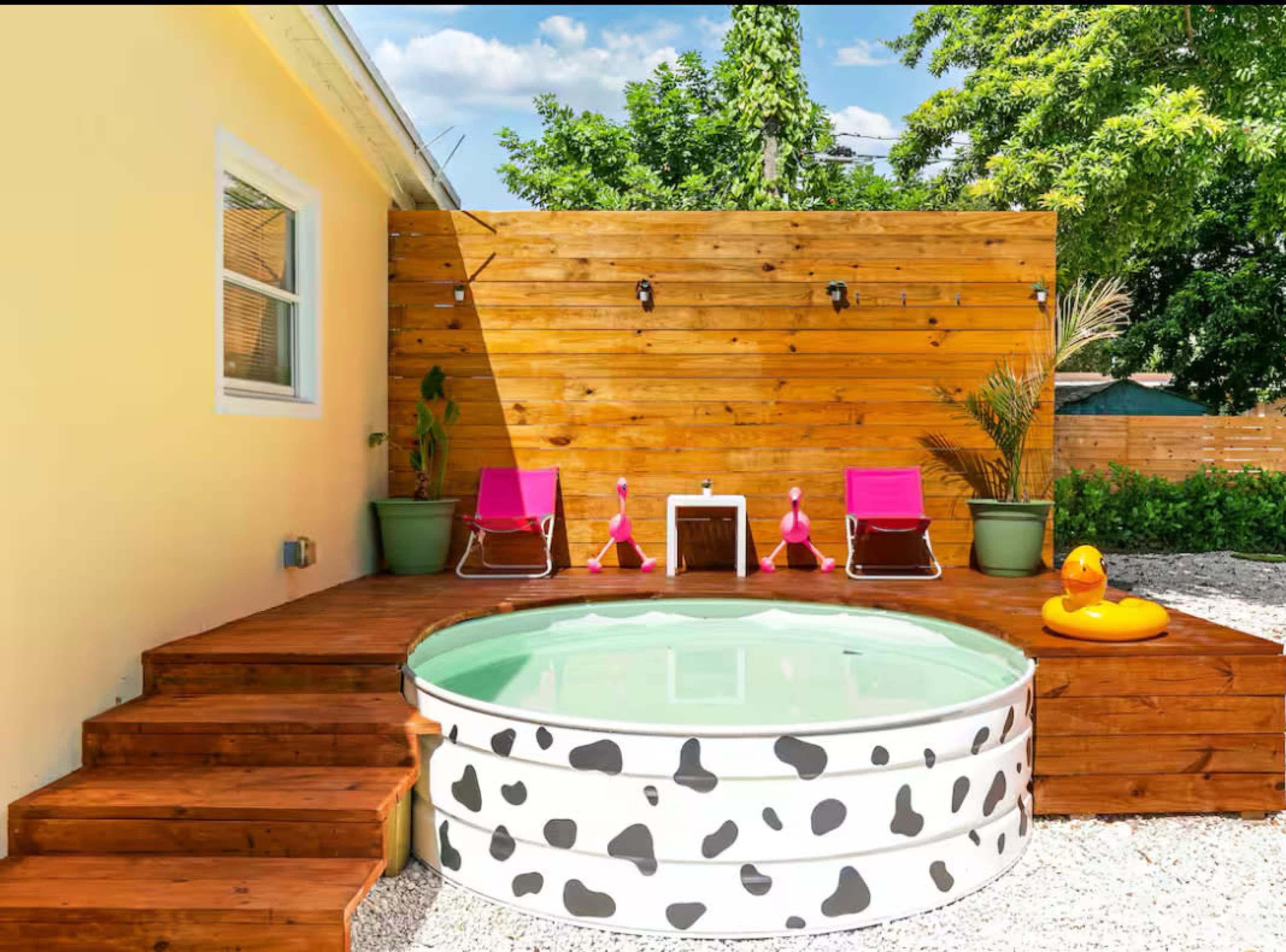 The image features a backyard with a circular, shallow pool surrounded by wooden decking, pink lounge chairs, and potted plants against a yellow wall and wooden fence.