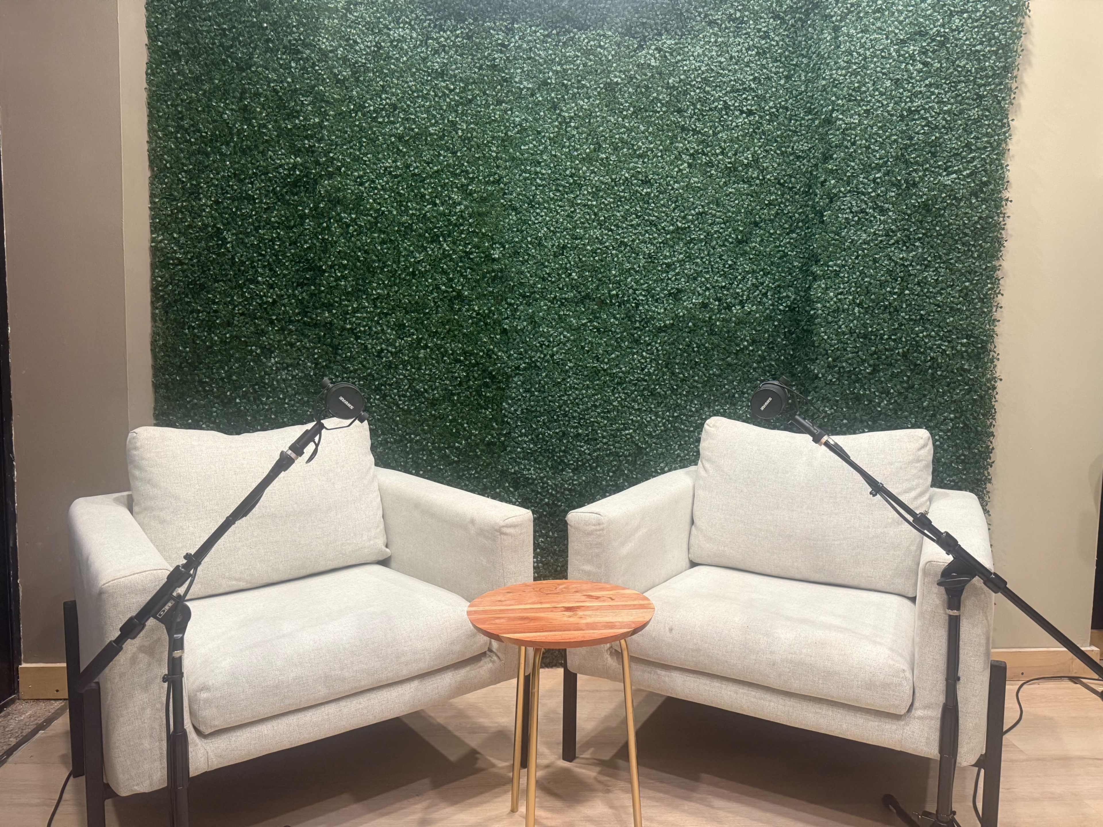 The image shows two light-colored armchairs facing each other with a small wooden table between them, set against a backdrop of green artificial foliage.