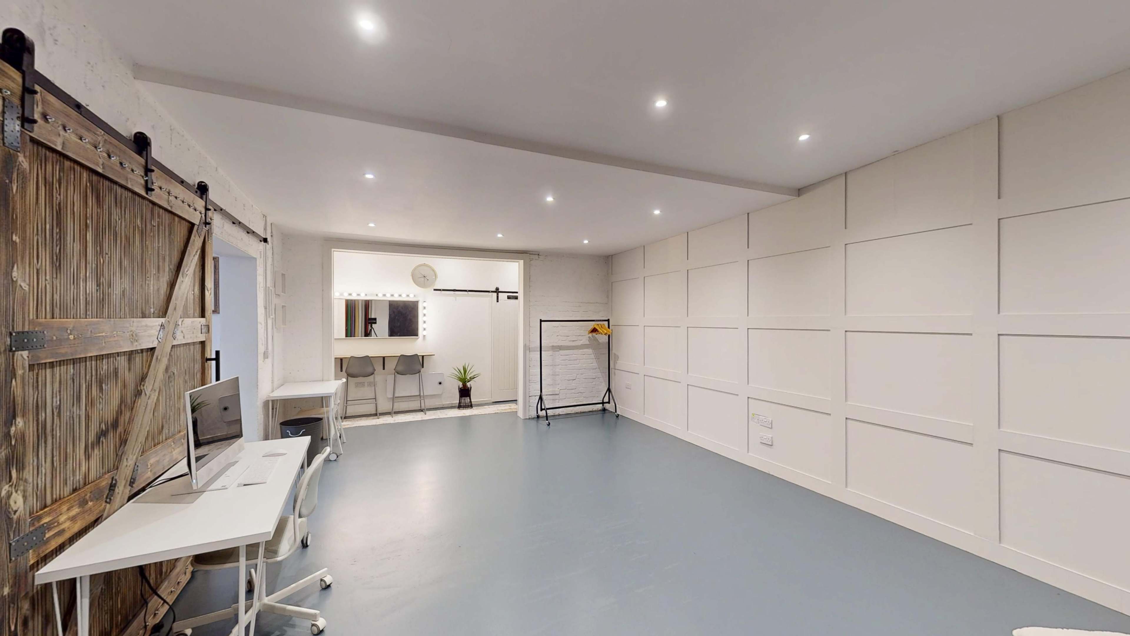 The image shows a spacious, well-lit room with a minimalist design featuring a desk and chair, a mirror with lighting, and a clothing rack against a white paneled wall.