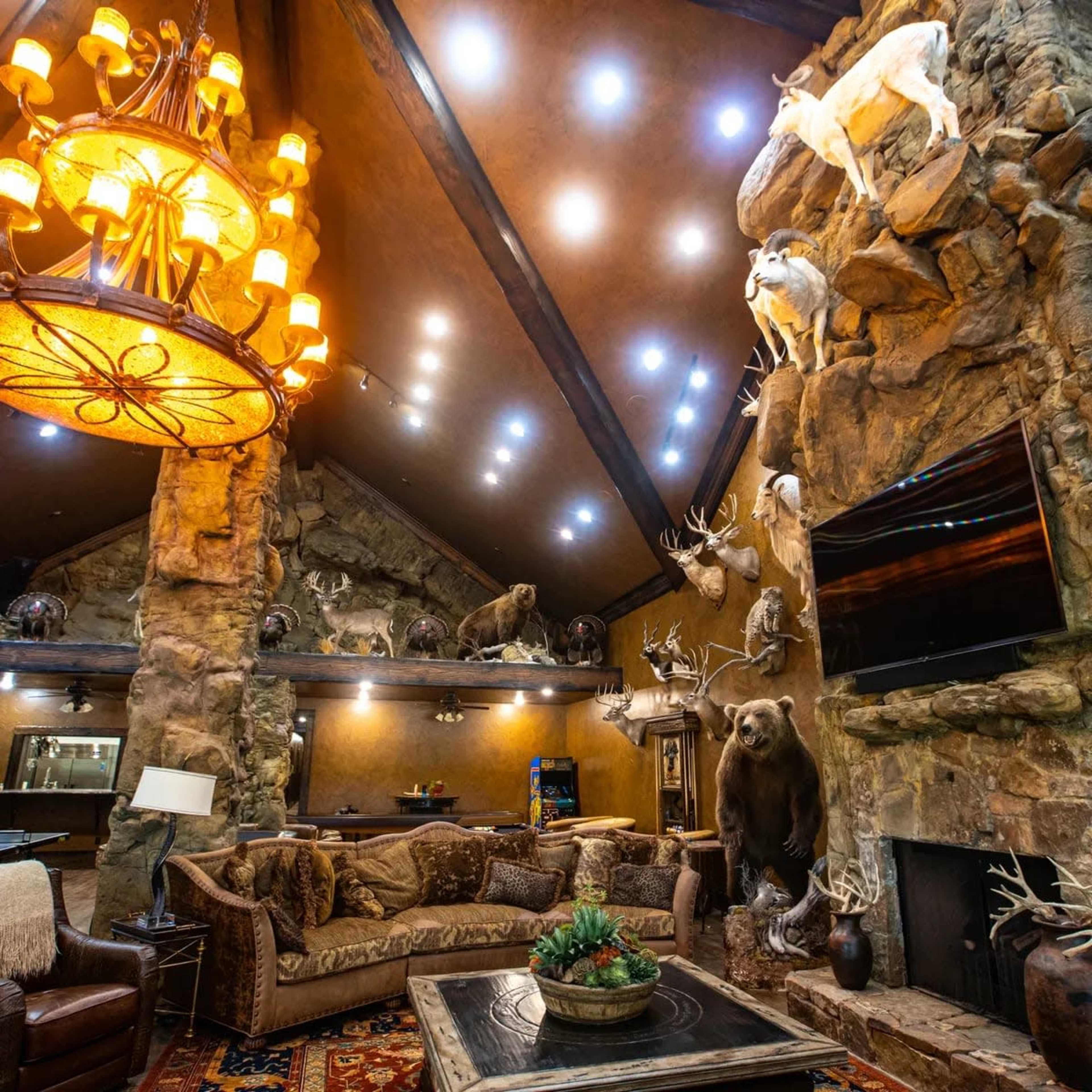 The image depicts a spacious lodge interior featuring a large stone fireplace, taxidermy animals mounted on the walls, and a cozy seating area with leather furniture.