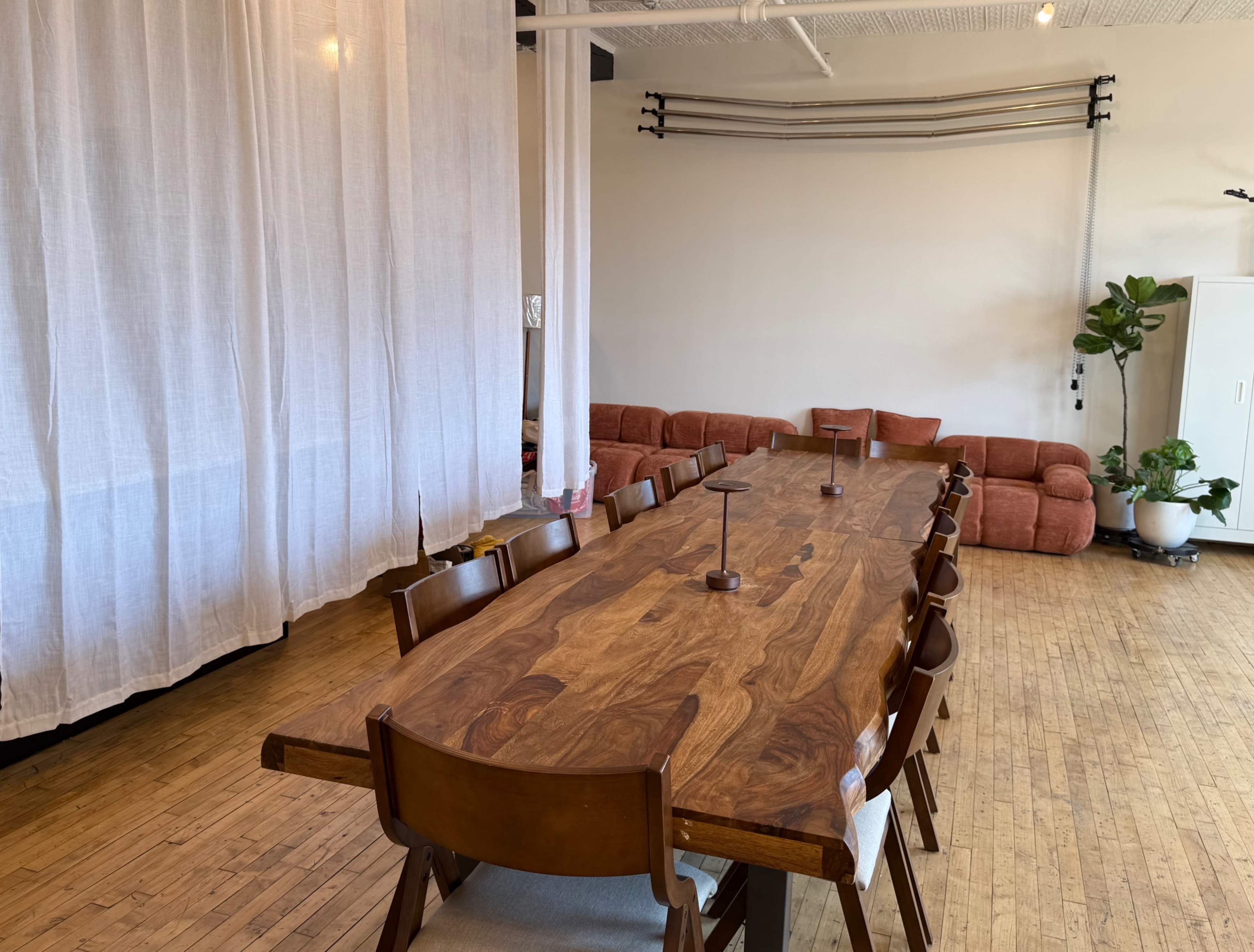 A long wooden table with eight chairs is set in a bright room featuring a white curtain, a green plant, and a sectional sofa along the wall.