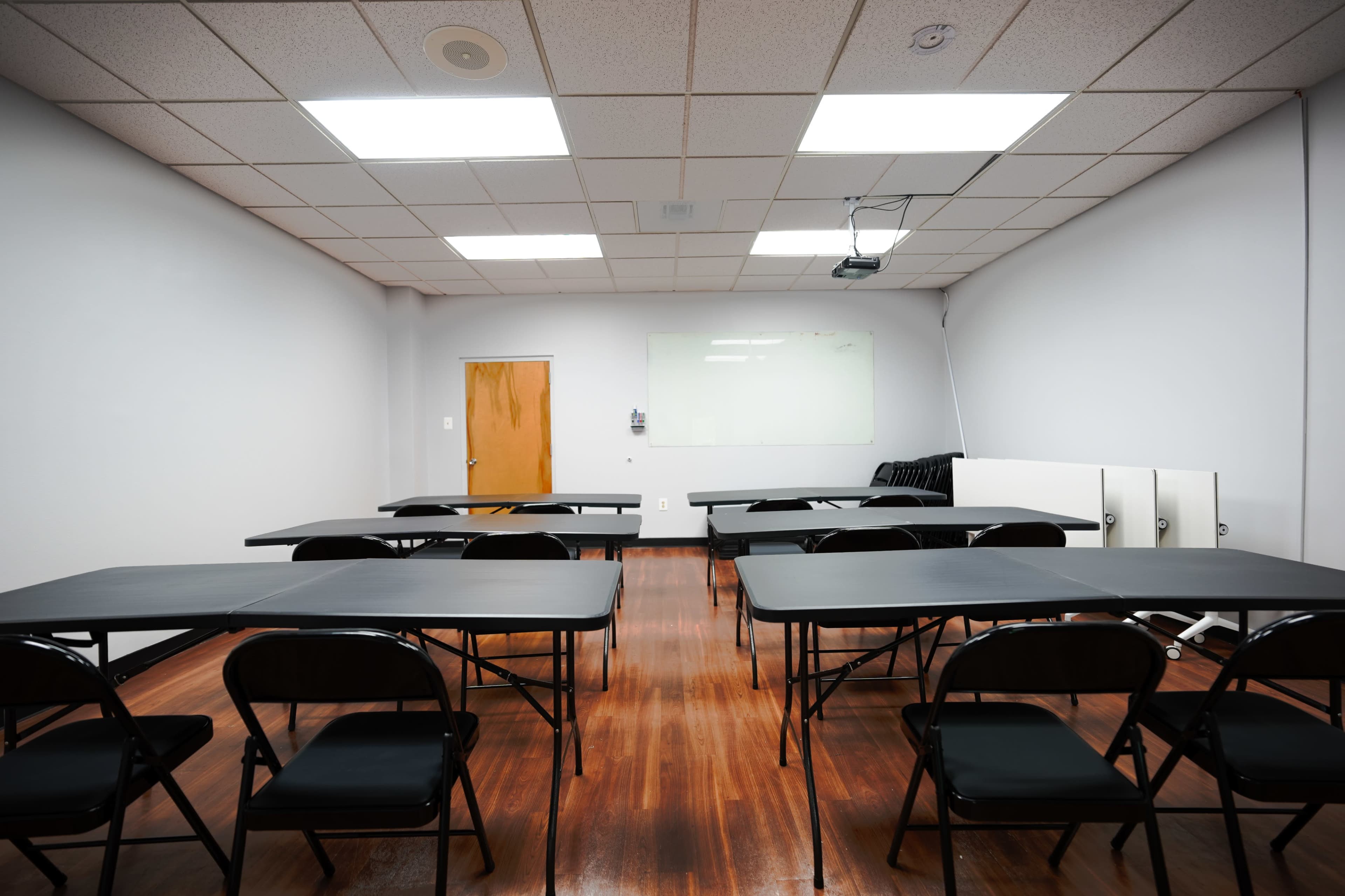 The image shows a sparsely furnished classroom with several black tables and chairs arranged in a symmetrical layout, a whiteboard, and a projector on the ceiling.