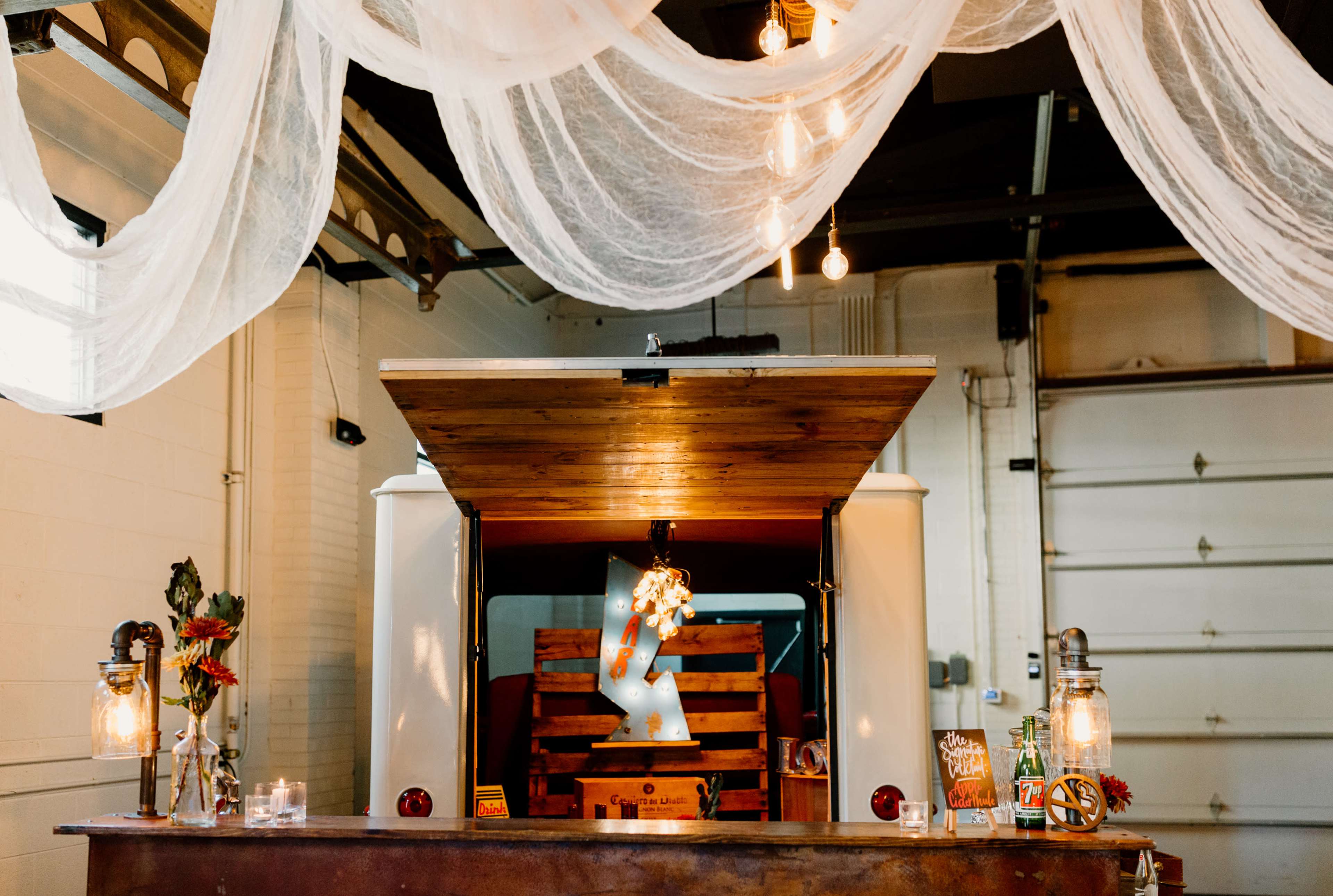 A rustic bar set up in a garage-like space, featuring a wooden counter, lanterns, and draped fabric overhead.