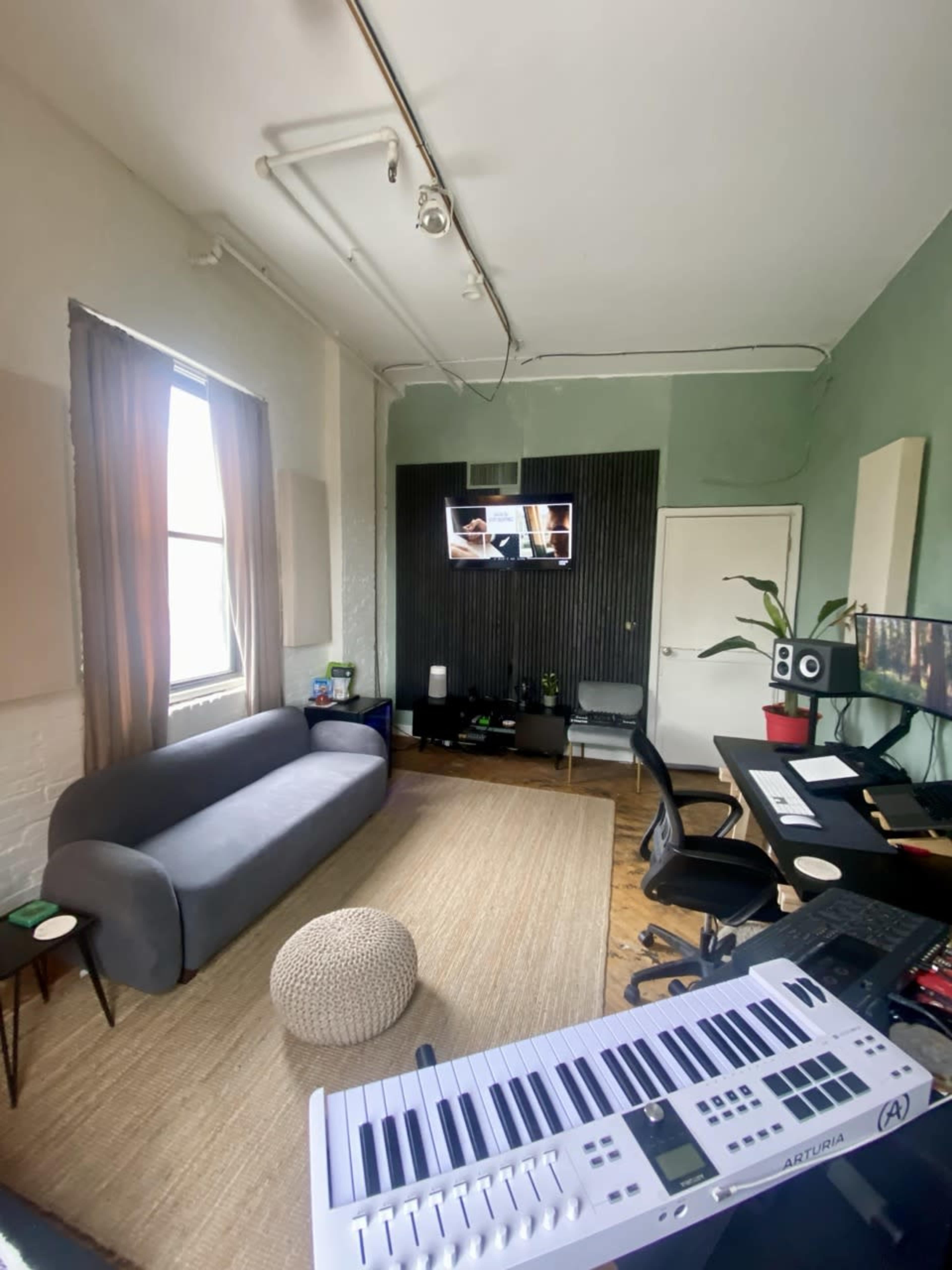 The image shows a cozy studio room featuring a gray sofa, a television mounted on a green accent wall, a desk with monitors, and a keyboard in the foreground.