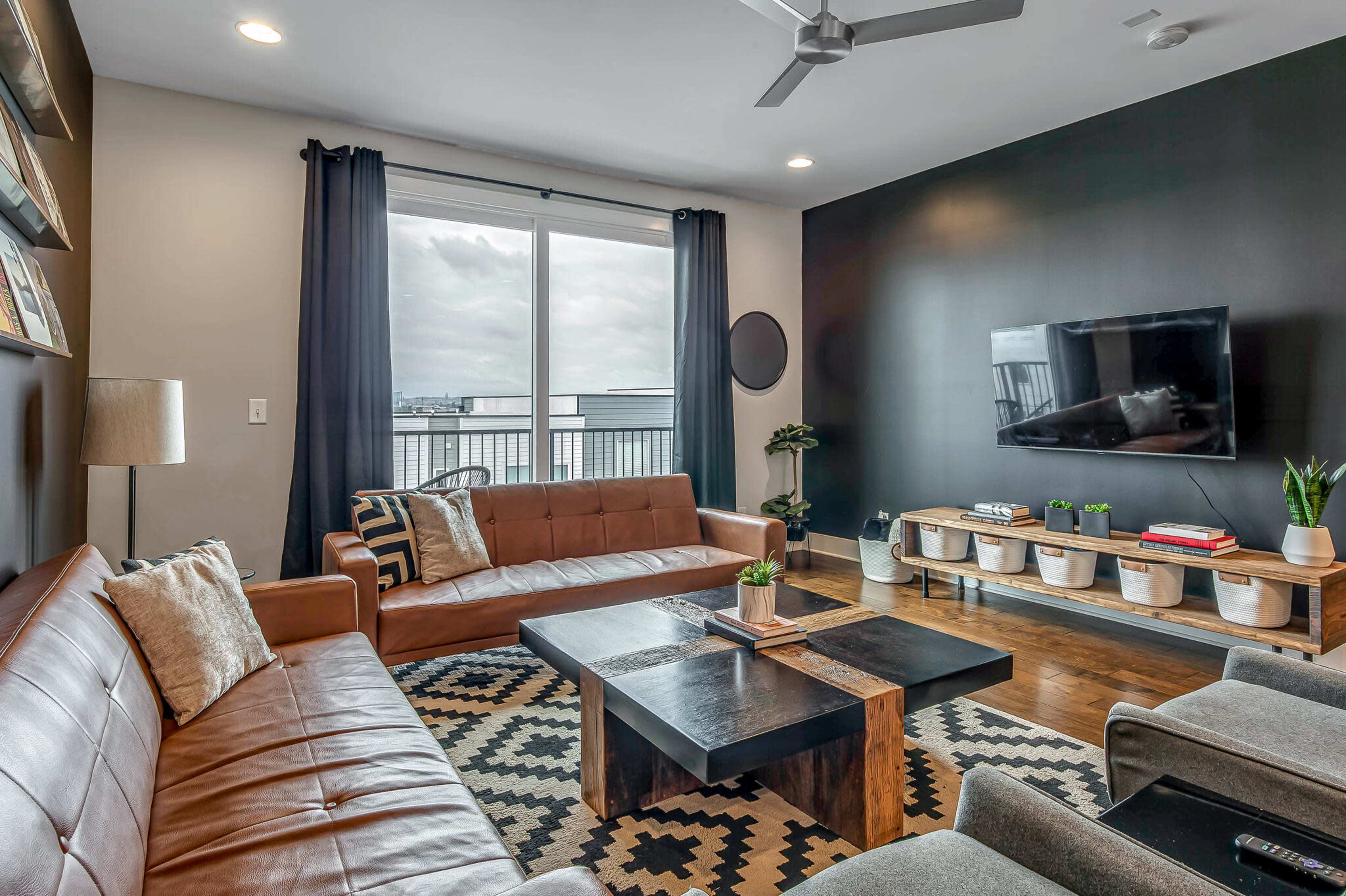 The image shows a modern living room featuring brown leather sofas, a dark wooden coffee table, a textured area rug, and a wall-mounted television, all complemented by large windows with black curtains.