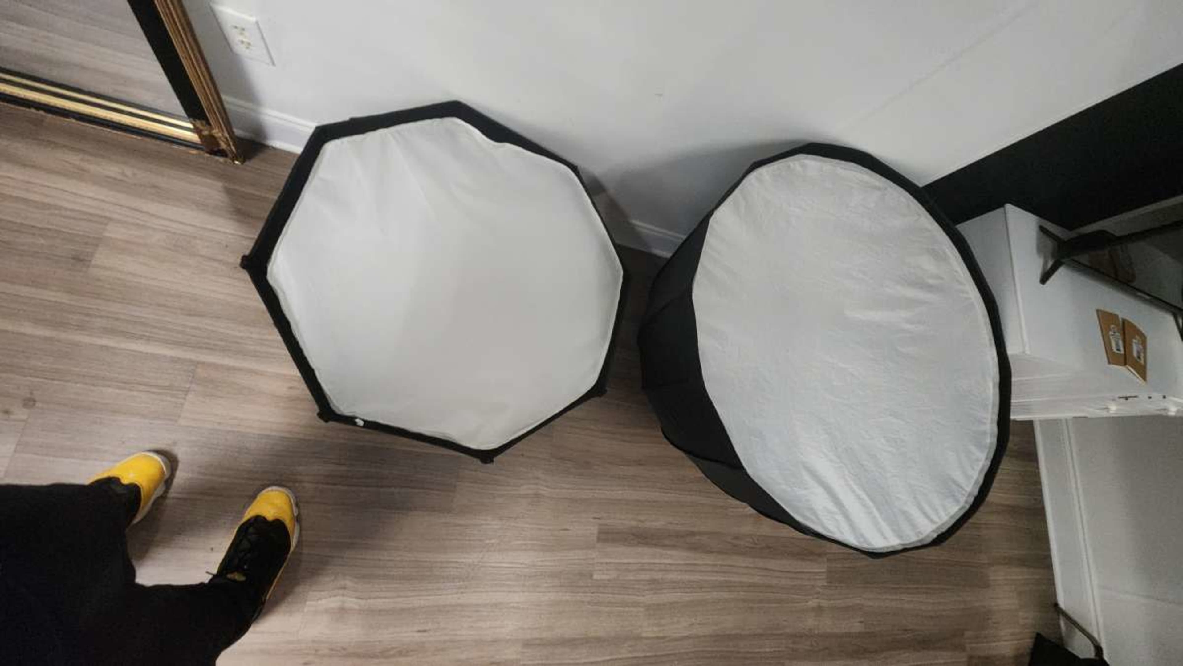 Two photography light reflectors, one octagonal and the other circular, are placed on a wooden floor next to a partially visible foot wearing yellow shoes.