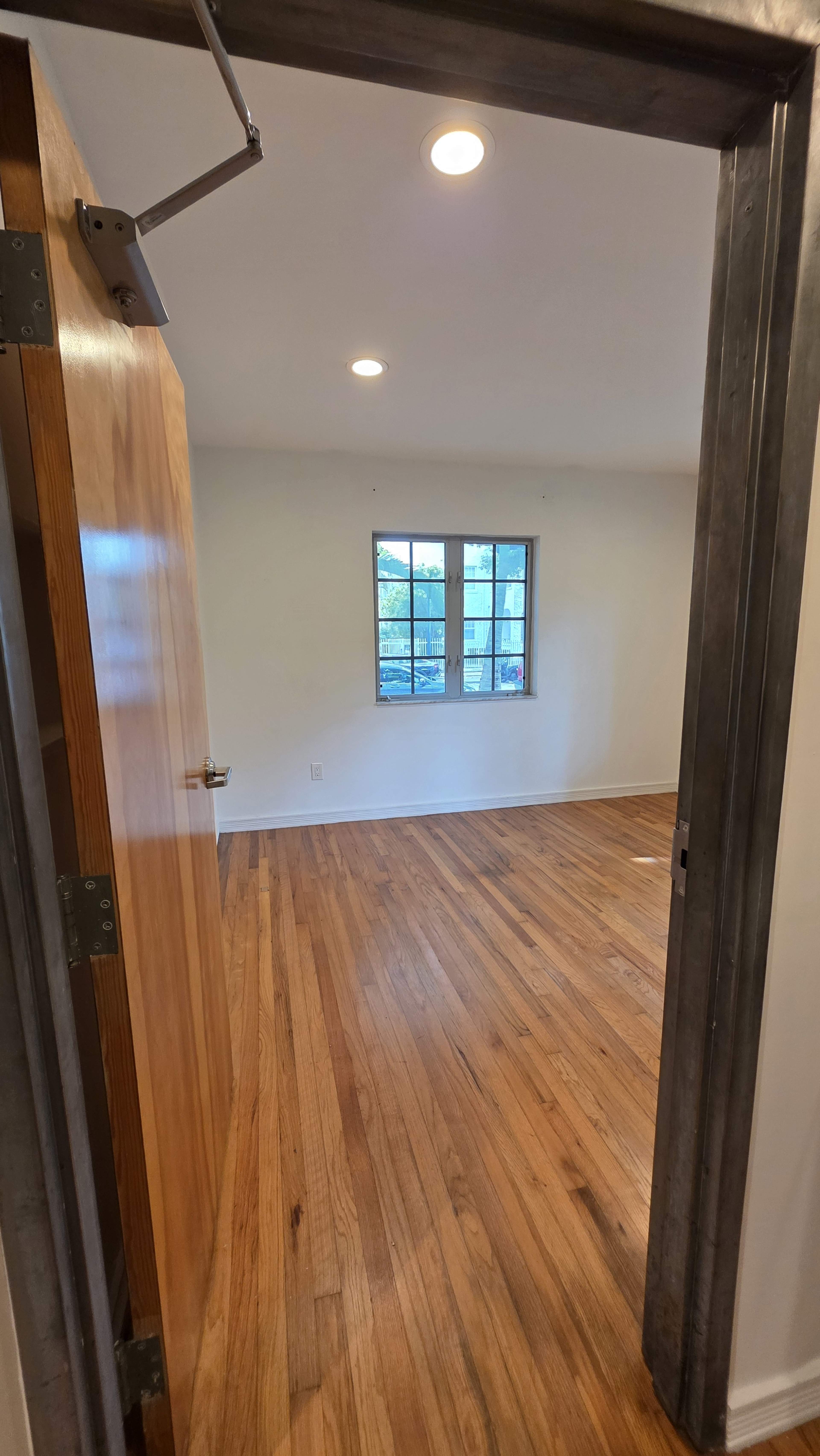 The image shows a vacant room with hardwood floors, a window, and an open door.