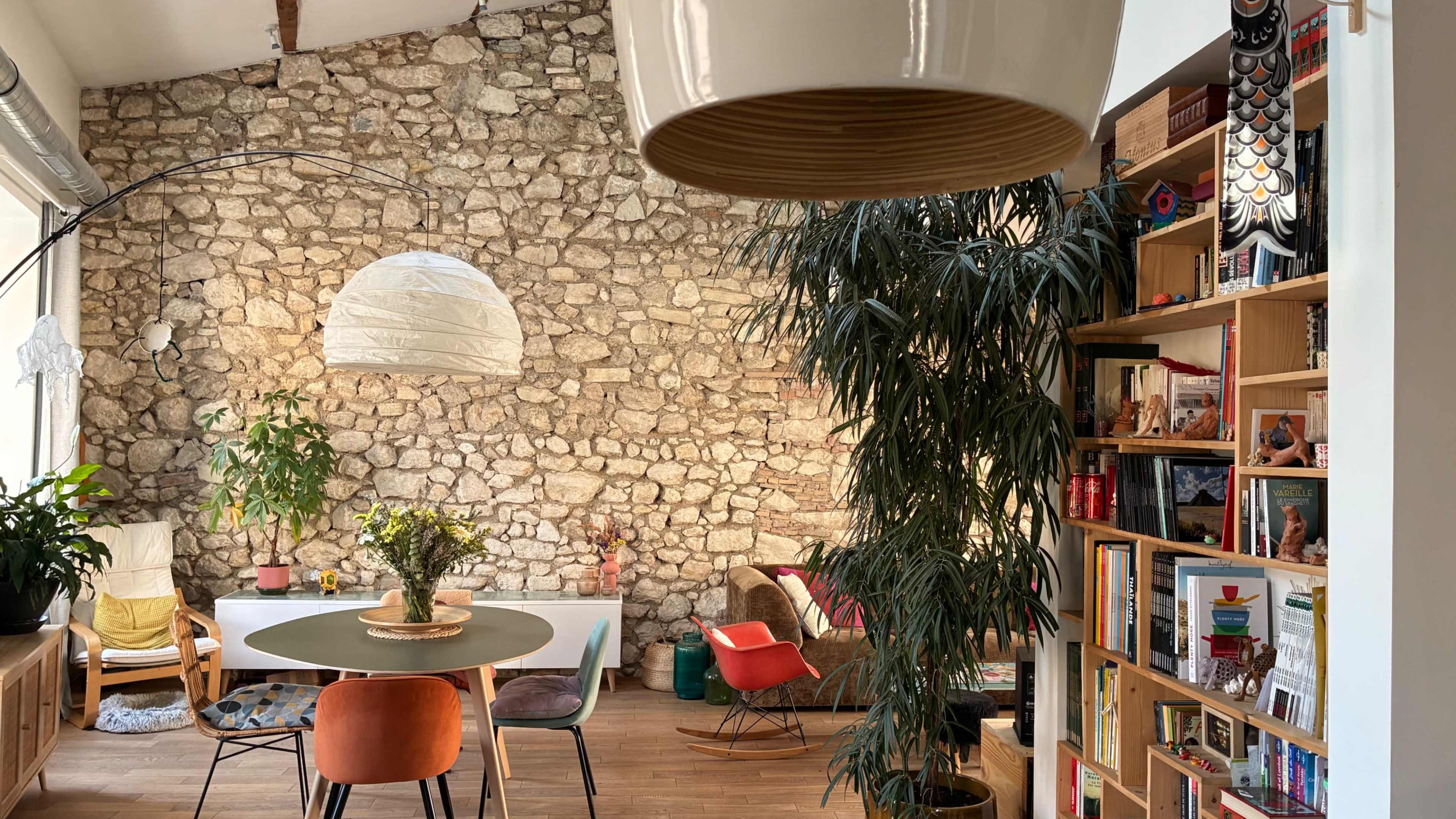 The scene shows a cozy living space with a stone wall, a round table surrounded by colorful chairs, and a bookshelf filled with various books and decorative items.