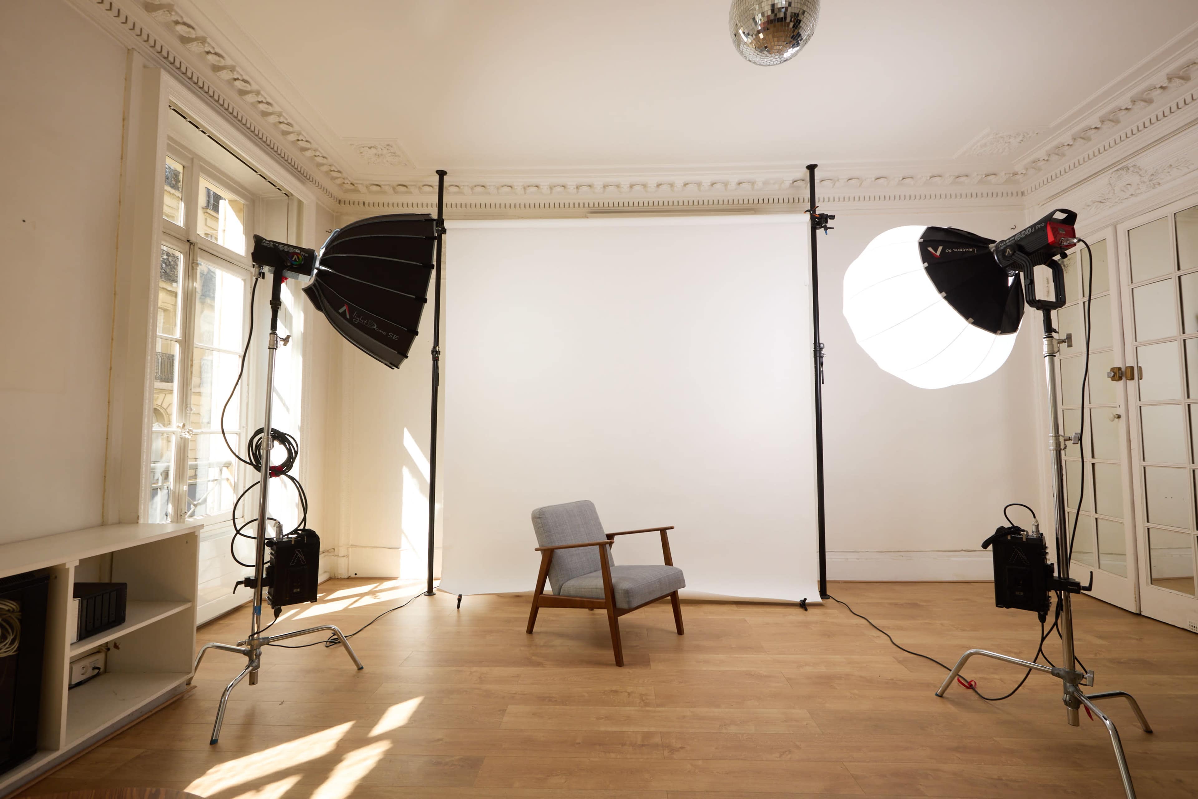 The image shows a photography studio with a gray armchair in front of a white backdrop, flanked by two softbox lights on stands.