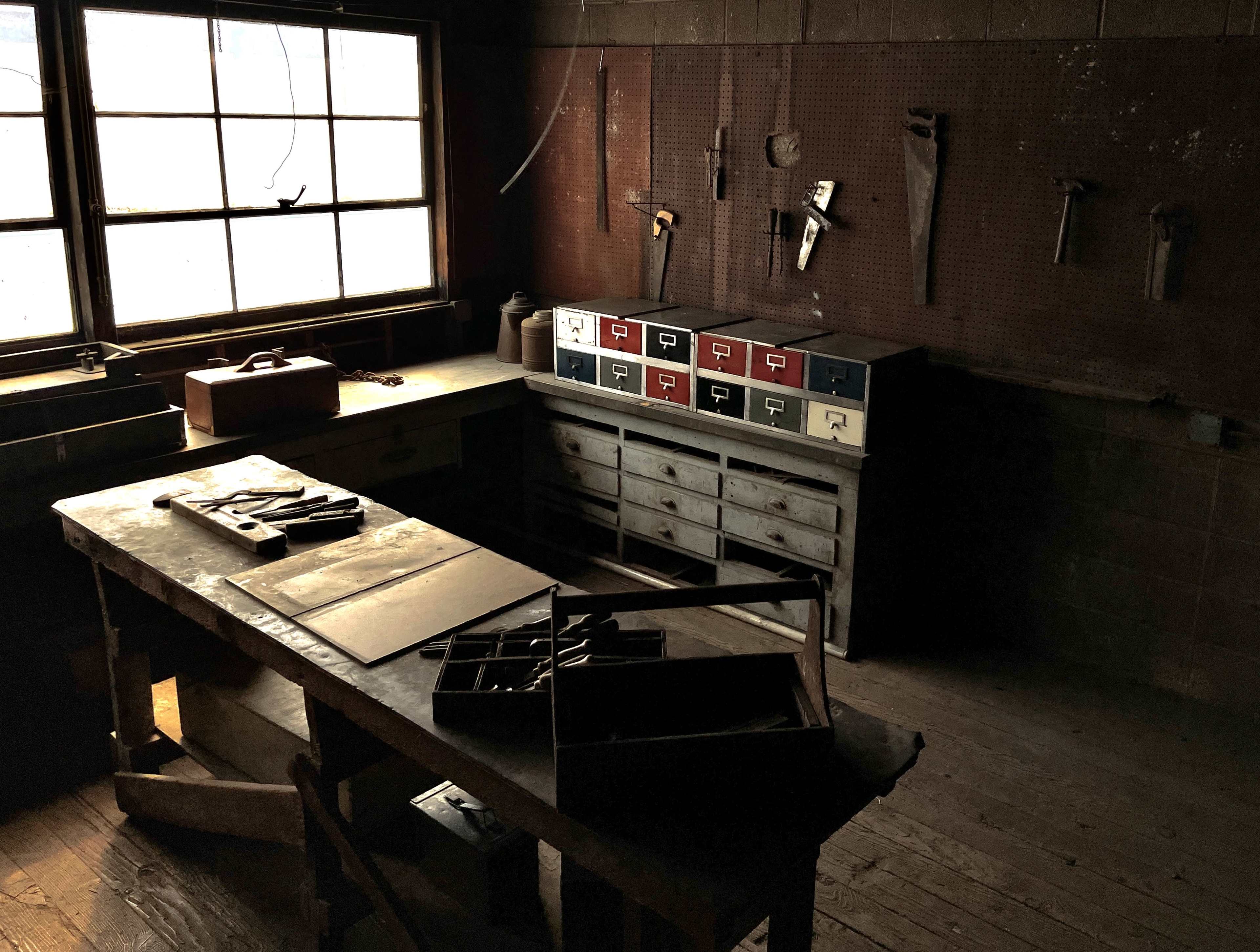 A dimly lit workshop with a wooden table, toolboxes, and various hand tools hanging on the wall.