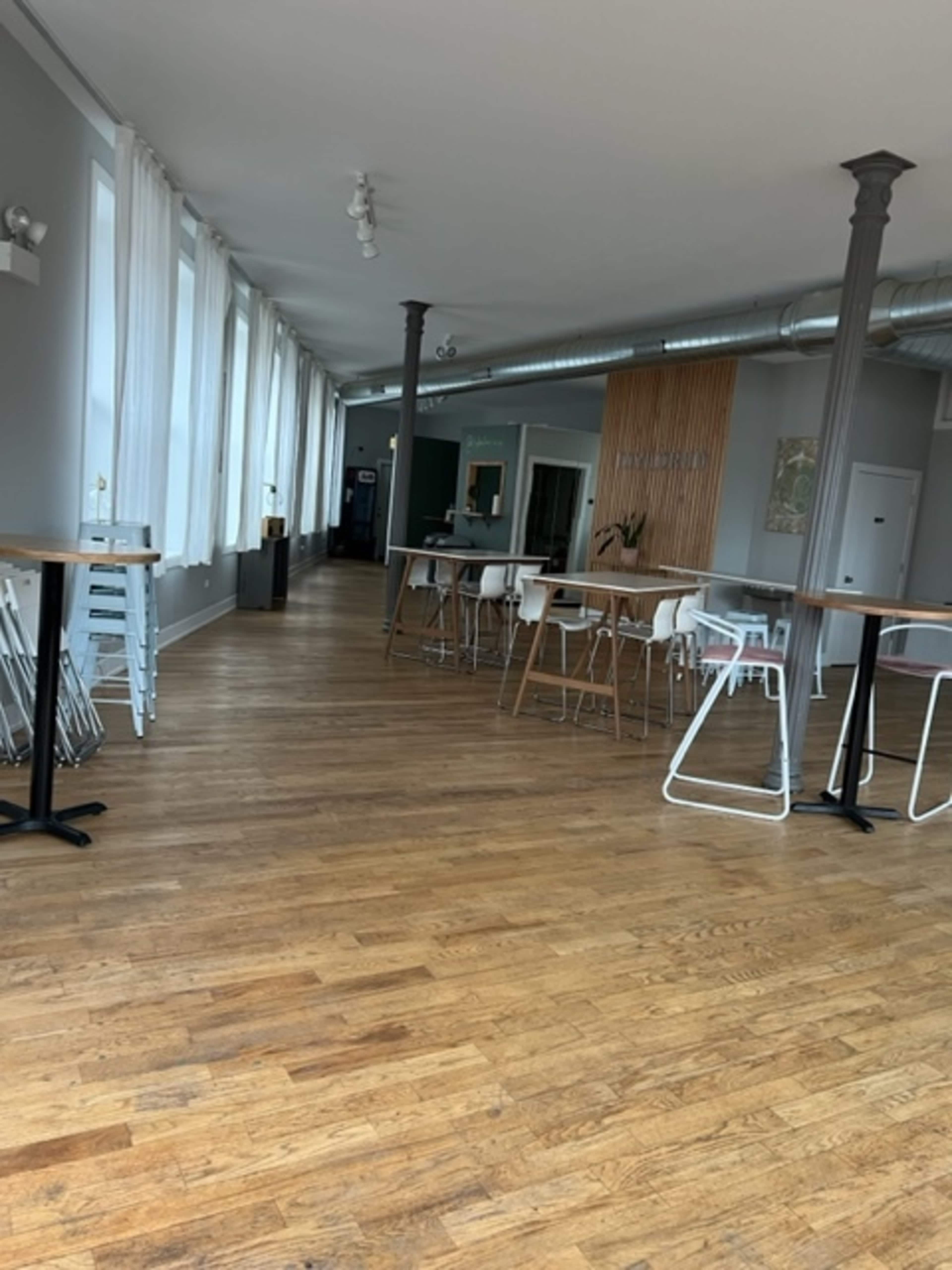 A spacious, modern interior with wooden floors and tall windows, featuring several tables and chairs arranged in a row.