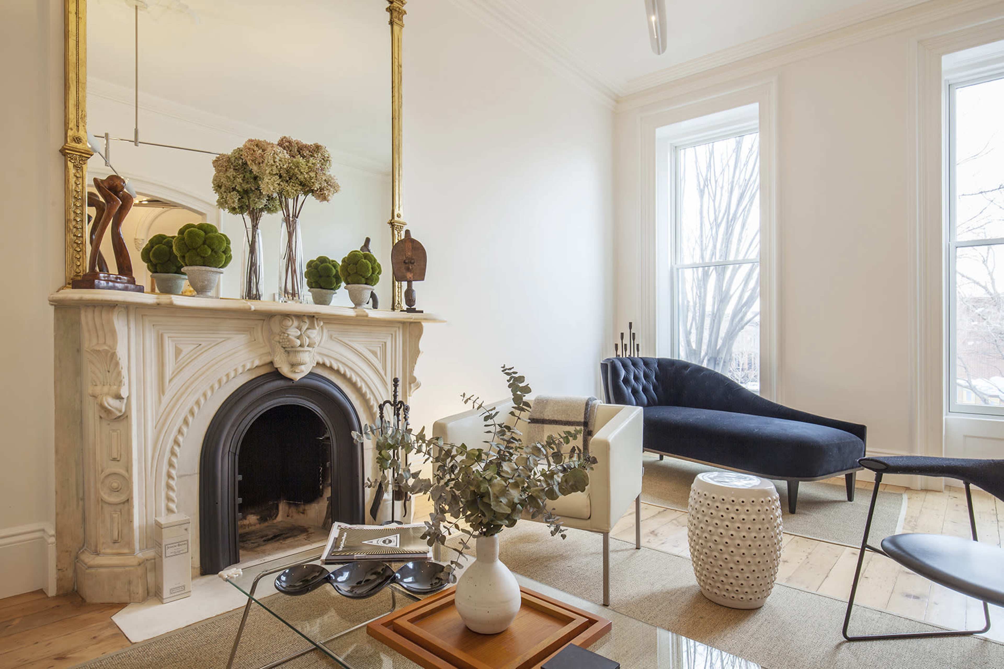 The image shows a living room with a marble fireplace, decorative plants, a dark blue sofa, and a coffee table on hardwood flooring.