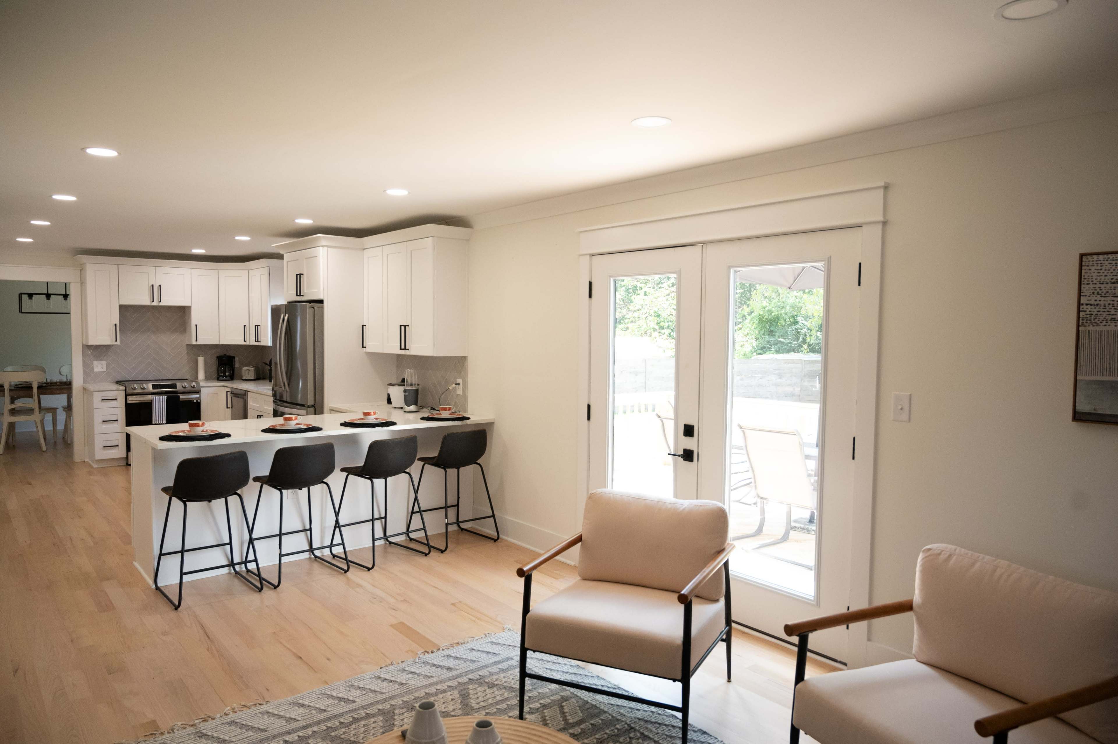 A modern kitchen with white cabinetry and a central island adjoins a living area with two armchairs and sliding glass doors leading to an outdoor patio.