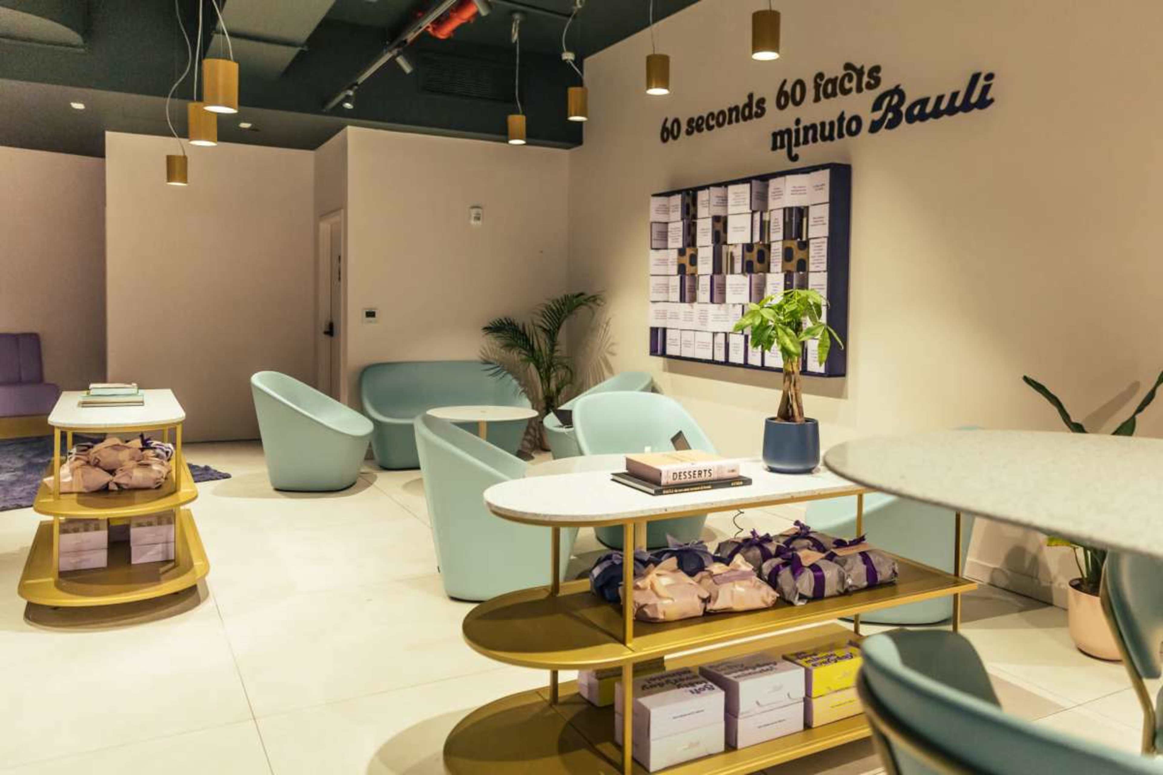 The image shows a modern interior space with colorful seating, decorative tables, and a wall featuring a calendar or schedule.