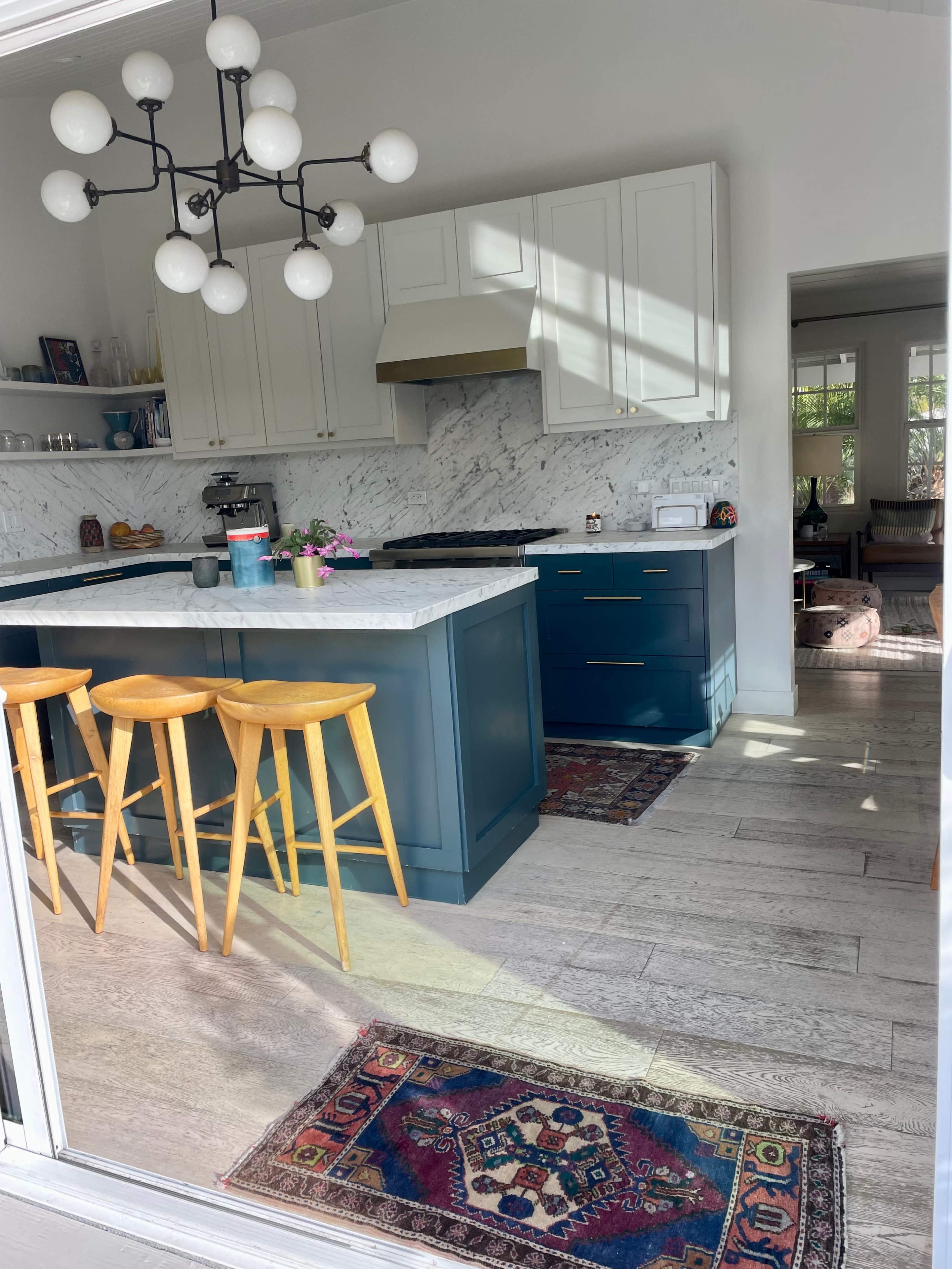A modern kitchen with a central island, bar stools, marble countertops, and a marble backsplash, illuminated by natural light.