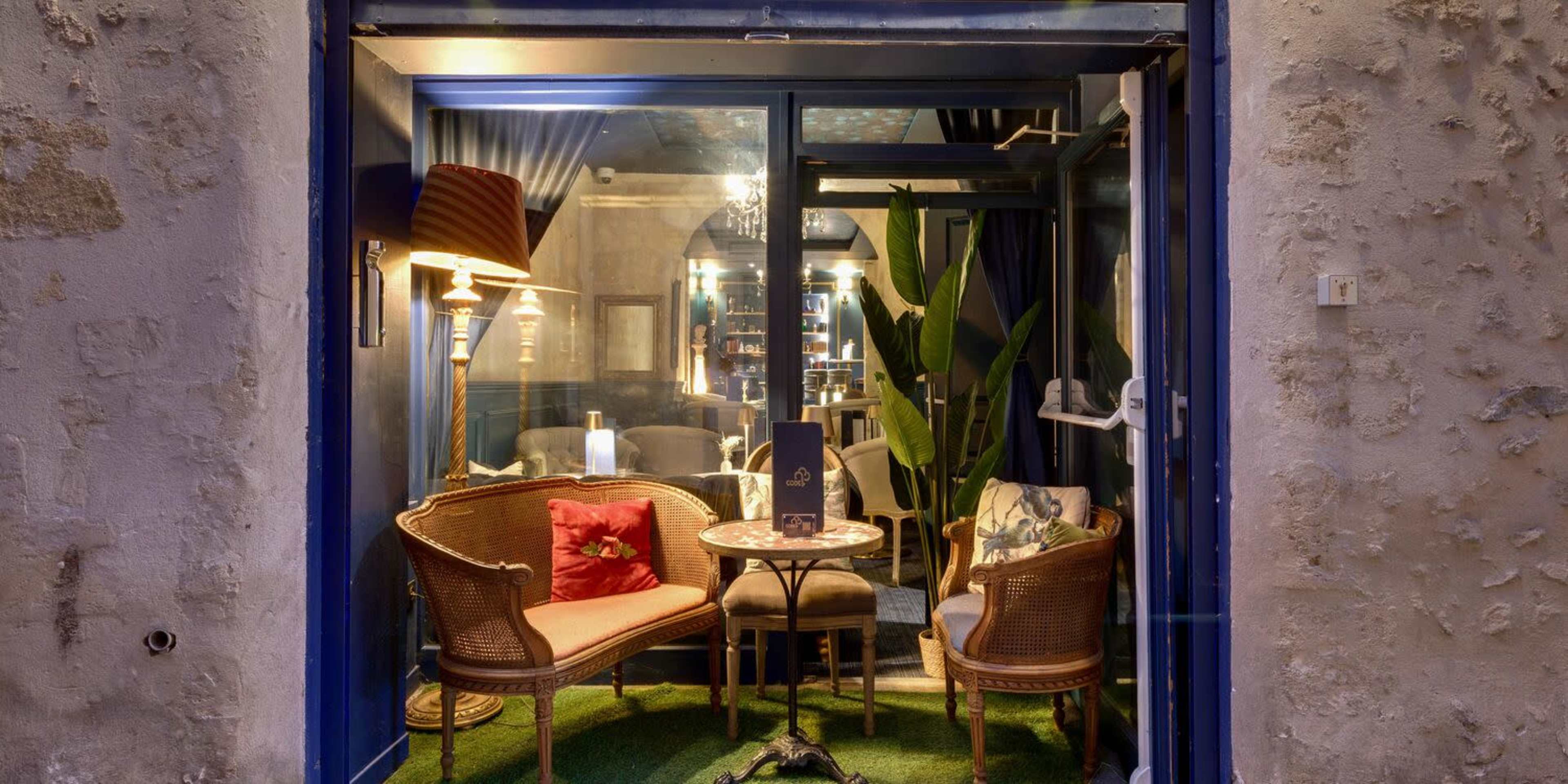The image shows a cozy café setting through a window, featuring two wicker chairs, a small table, and decorative plants.