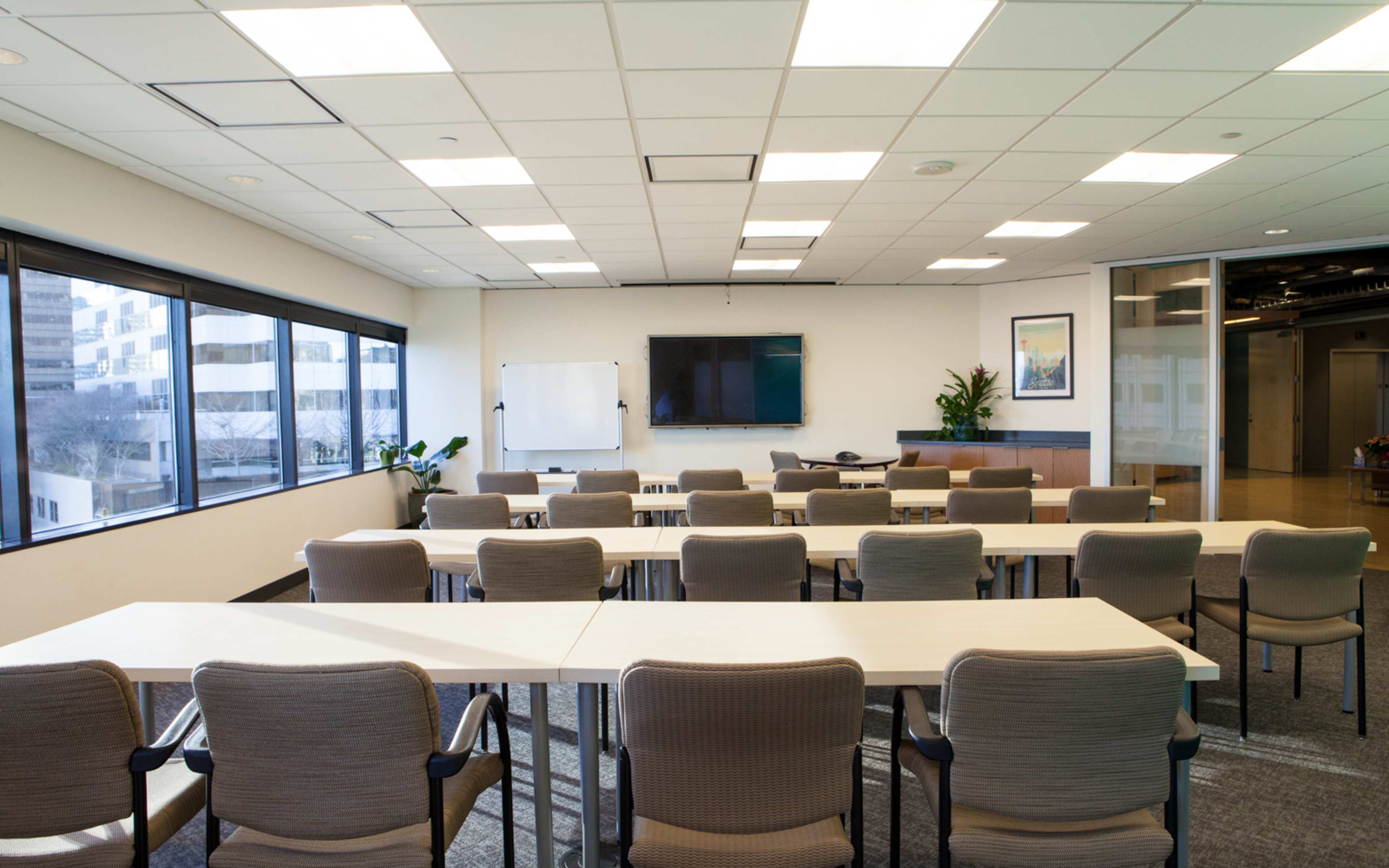 A well-lit conference room with rows of tables and chairs, a presentation screen, and large windows overlooking a cityscape.