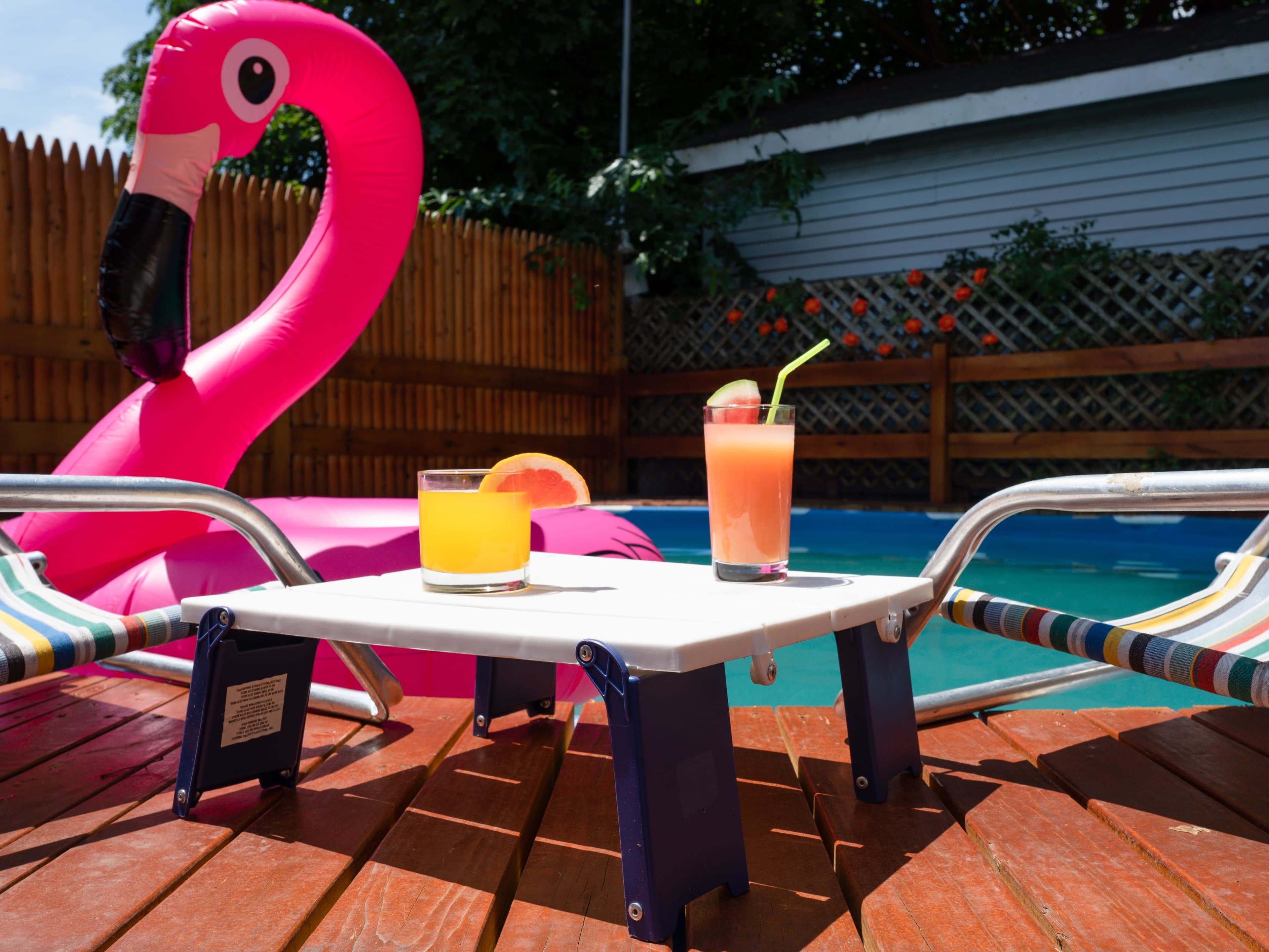 A table holds two colorful cocktails beside a pink inflatable flamingo by a swimming pool.