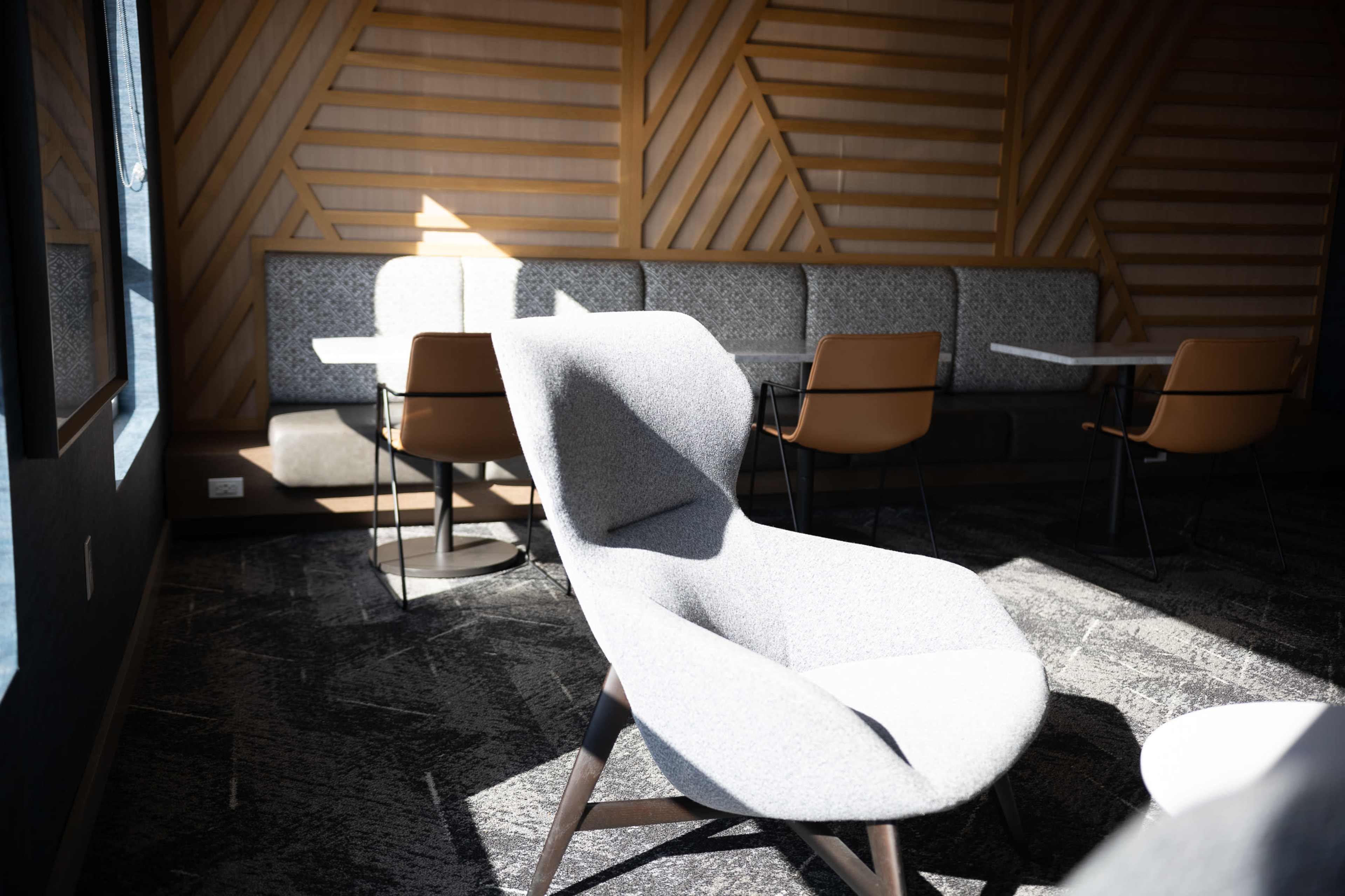 A gray lounge chair is positioned in a well-lit corner of a modern seating area with textured wooden walls and a row of tables and chairs in the background.