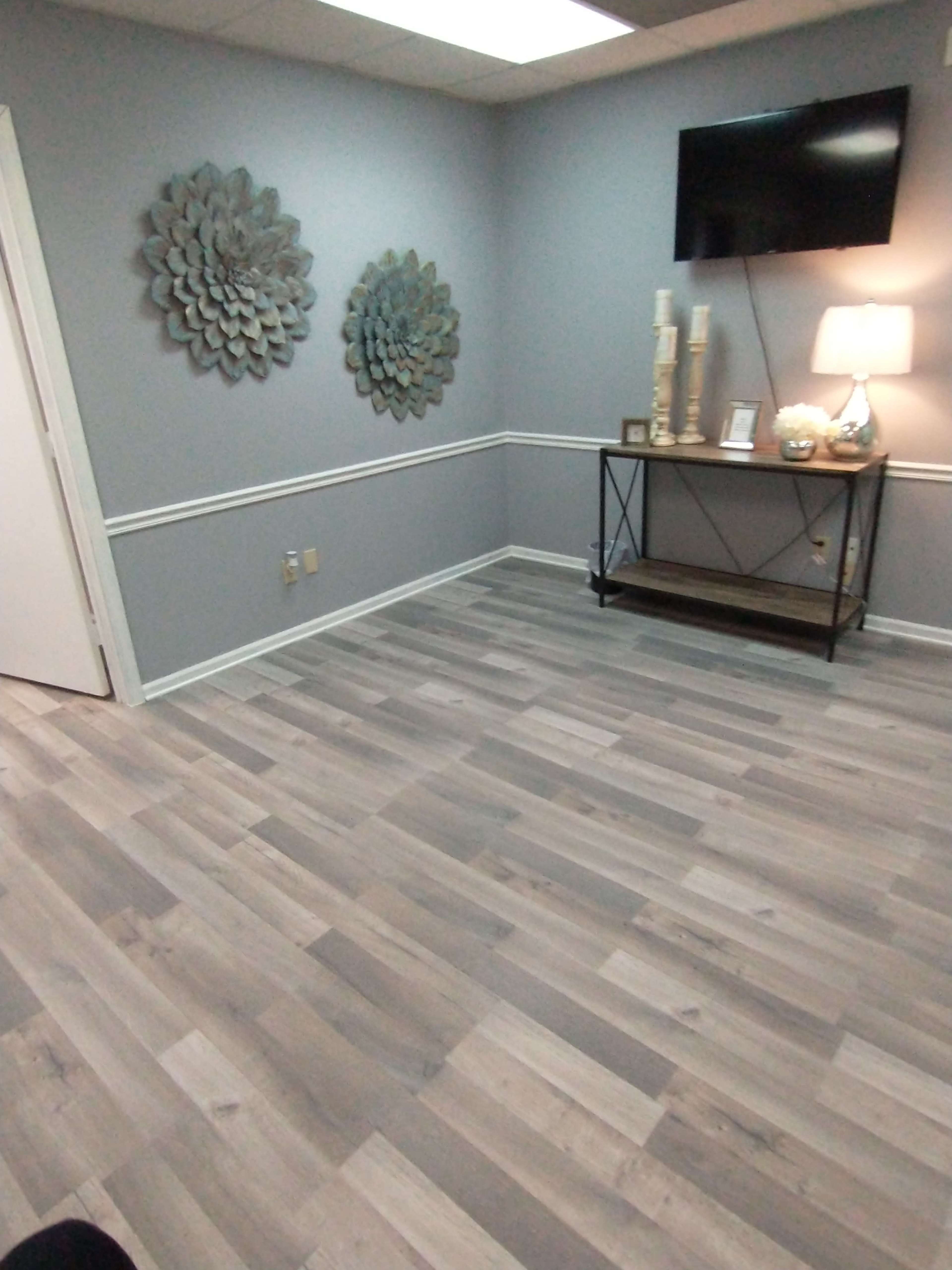 The room features a light gray wall, two decorative wall pieces, a TV mounted on the wall, and a lamp on a black console table.