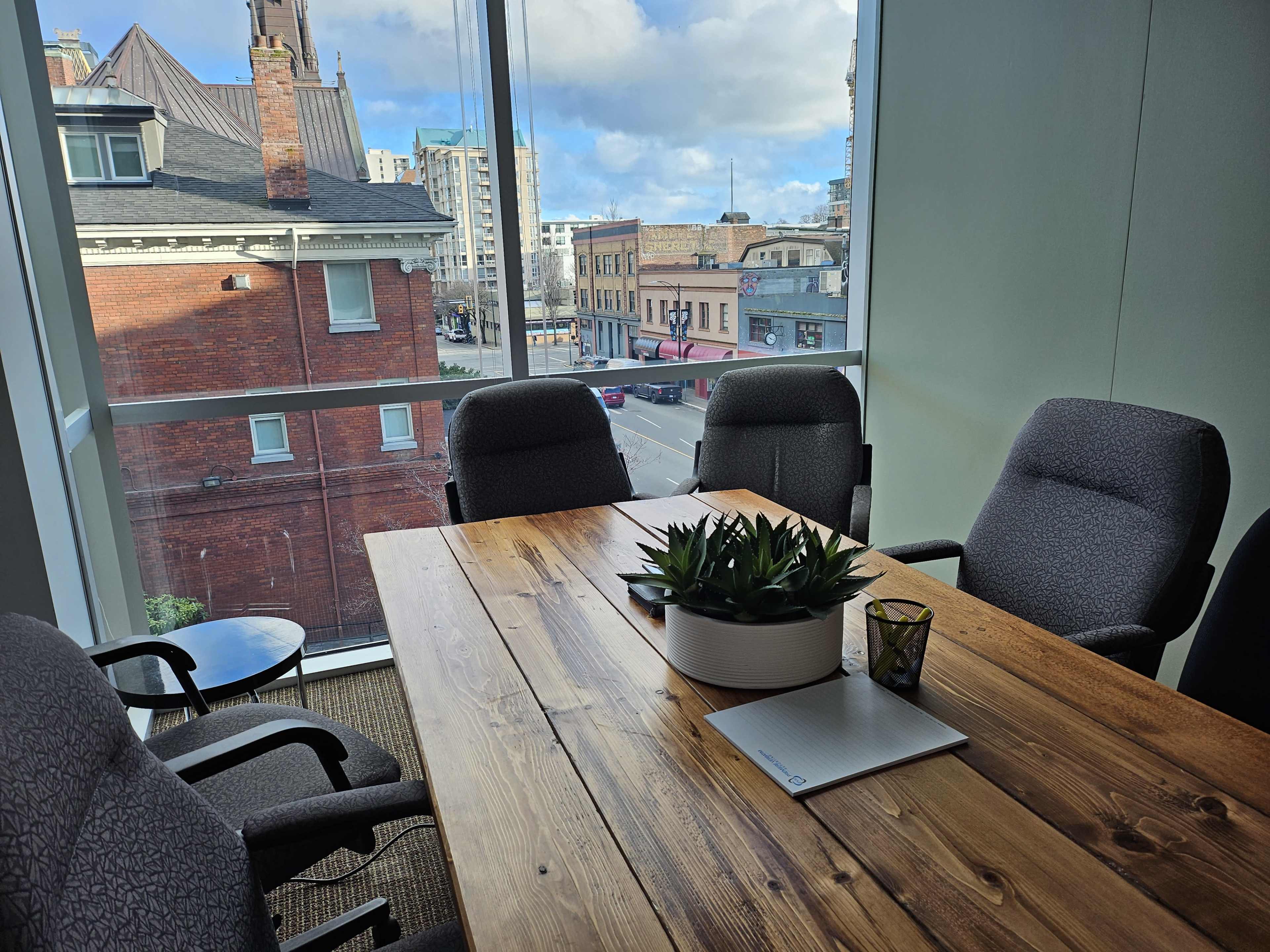 A meeting room features a large wooden table surrounded by chairs, with a view of a city street through large windows.
